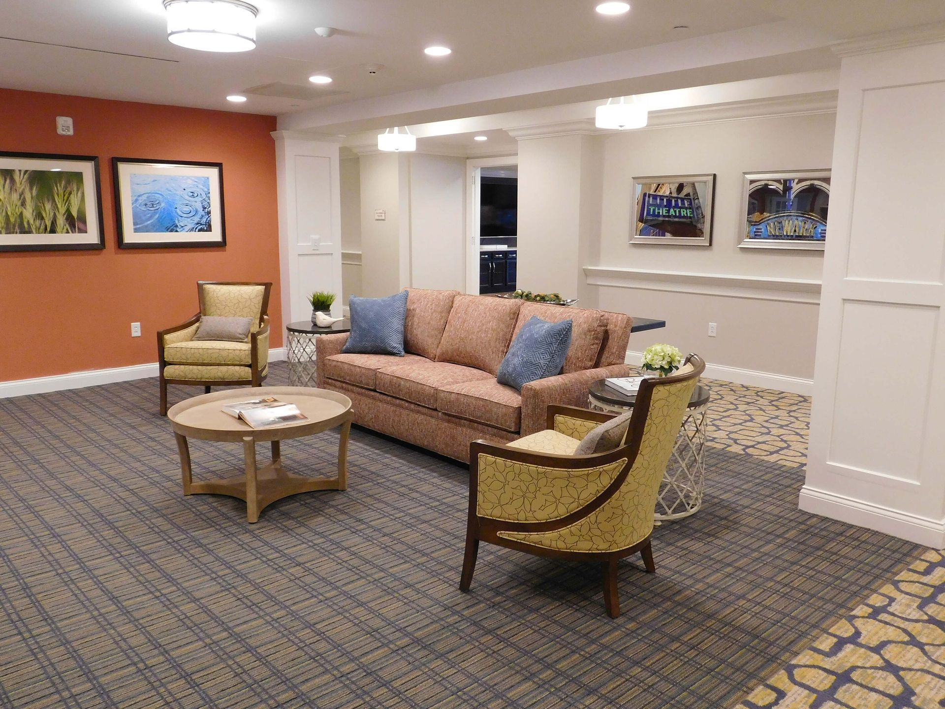 Modern lounge with tan sofa, yellow chairs, round tables, and framed art on white and orange walls.