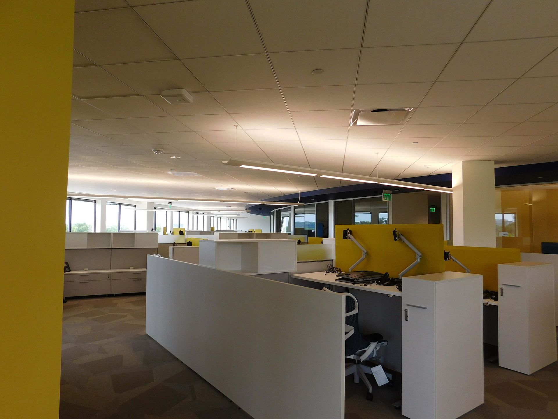 Open office cubicles with yellow walls and white desks under fluorescent lights