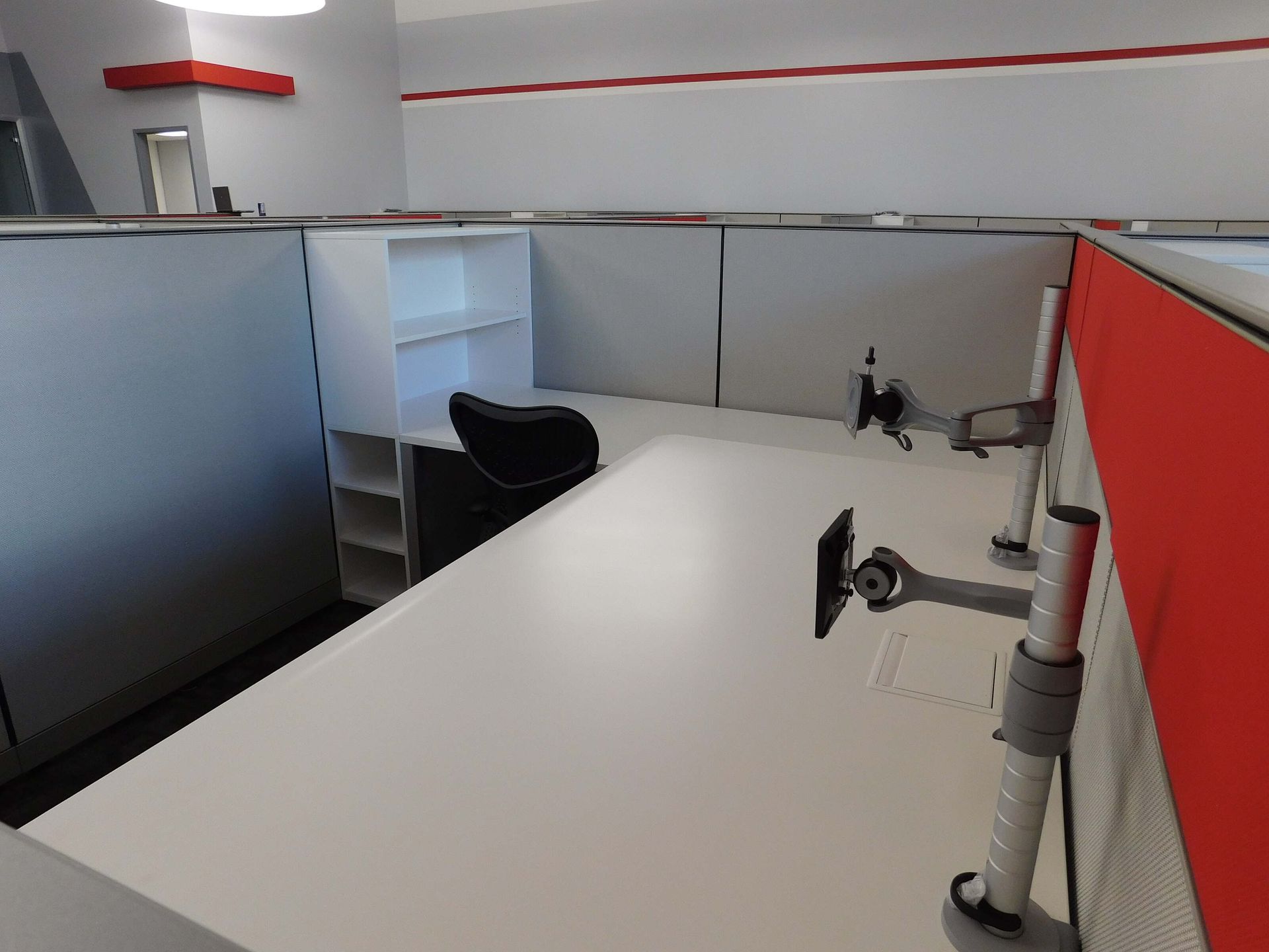 Empty office cubicle with white desk, black chair, and gray partitions with red trim.