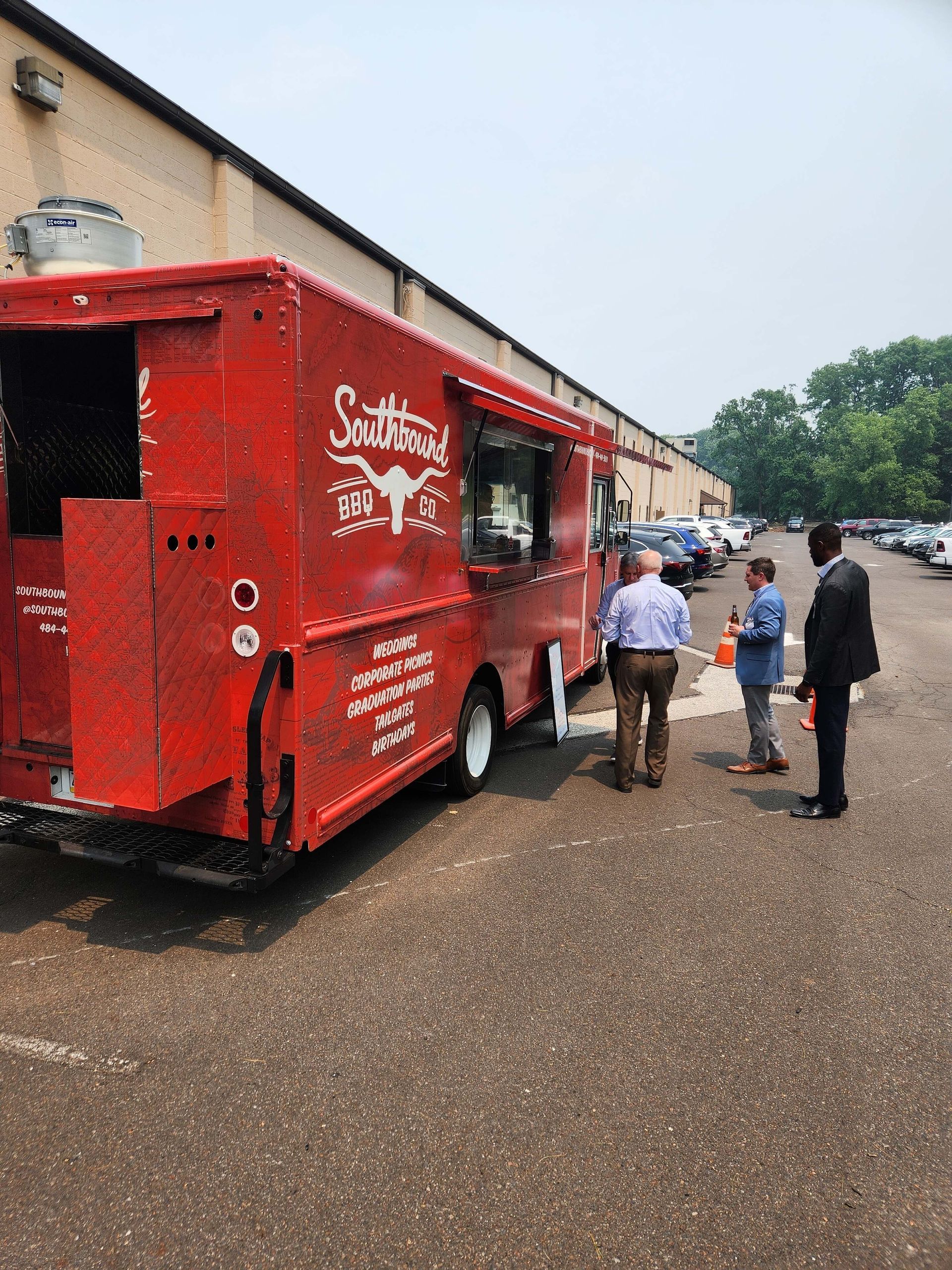Red food truck serving people outside a warehouse on a gravel lot
