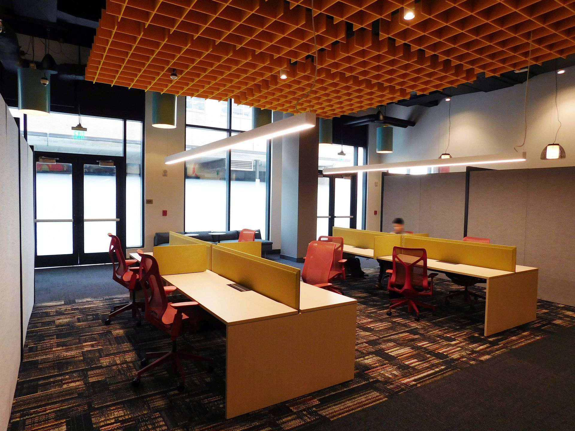 Modern office lounge with red chairs, yellow booths, and long tables under a wood lattice ceiling.