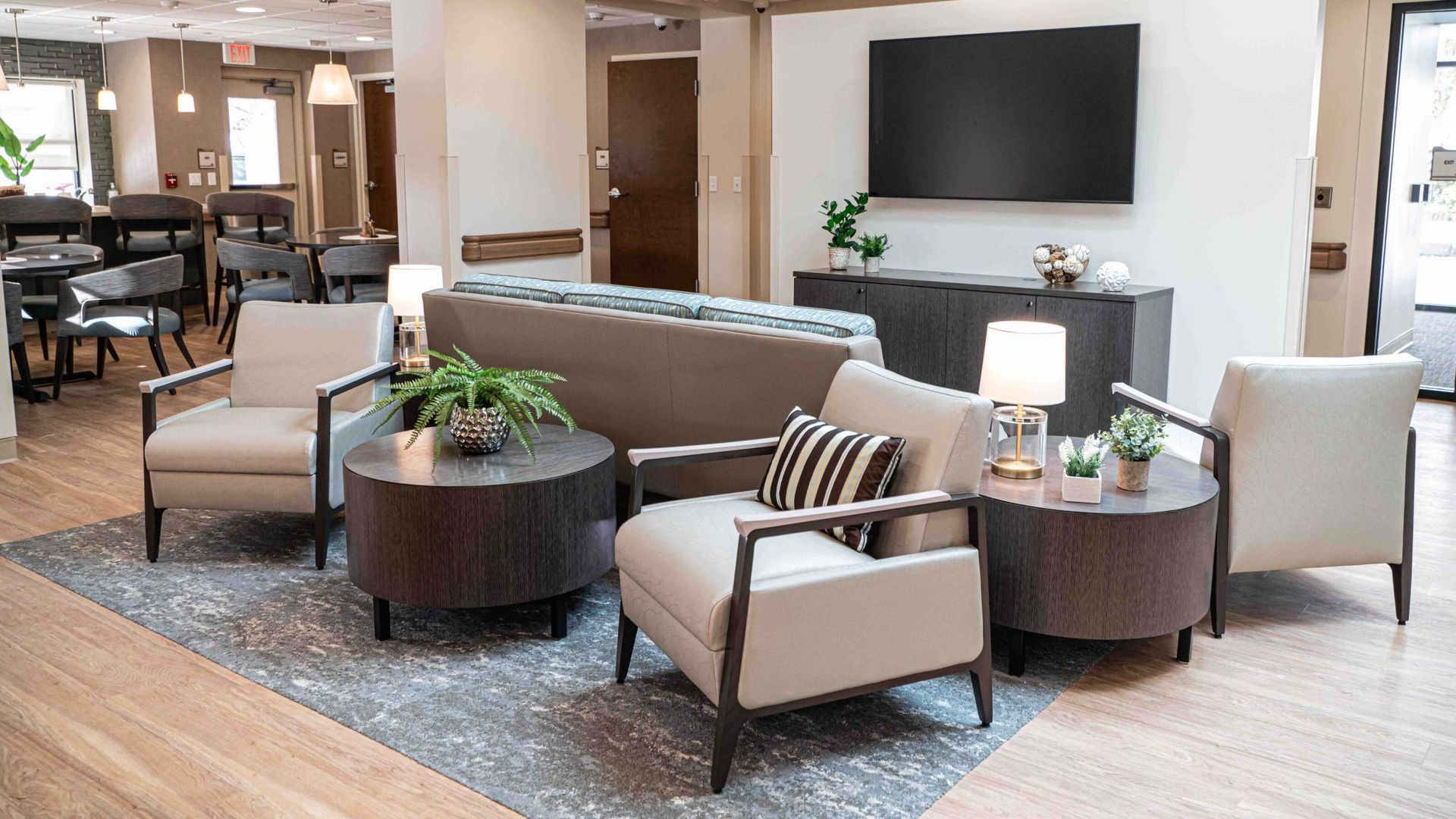 Modern lobby seating area with beige chairs, round tables, plants, and a wall-mounted TV