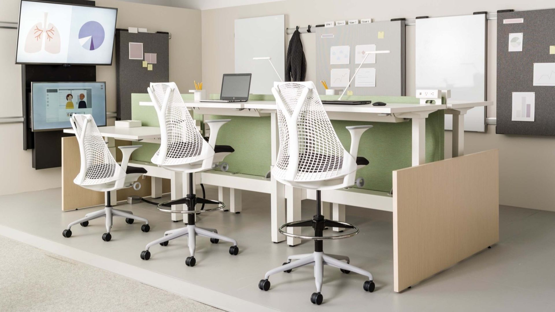 Modern office with white chairs, shared desks, laptops, and wall-mounted screens in a bright workspace