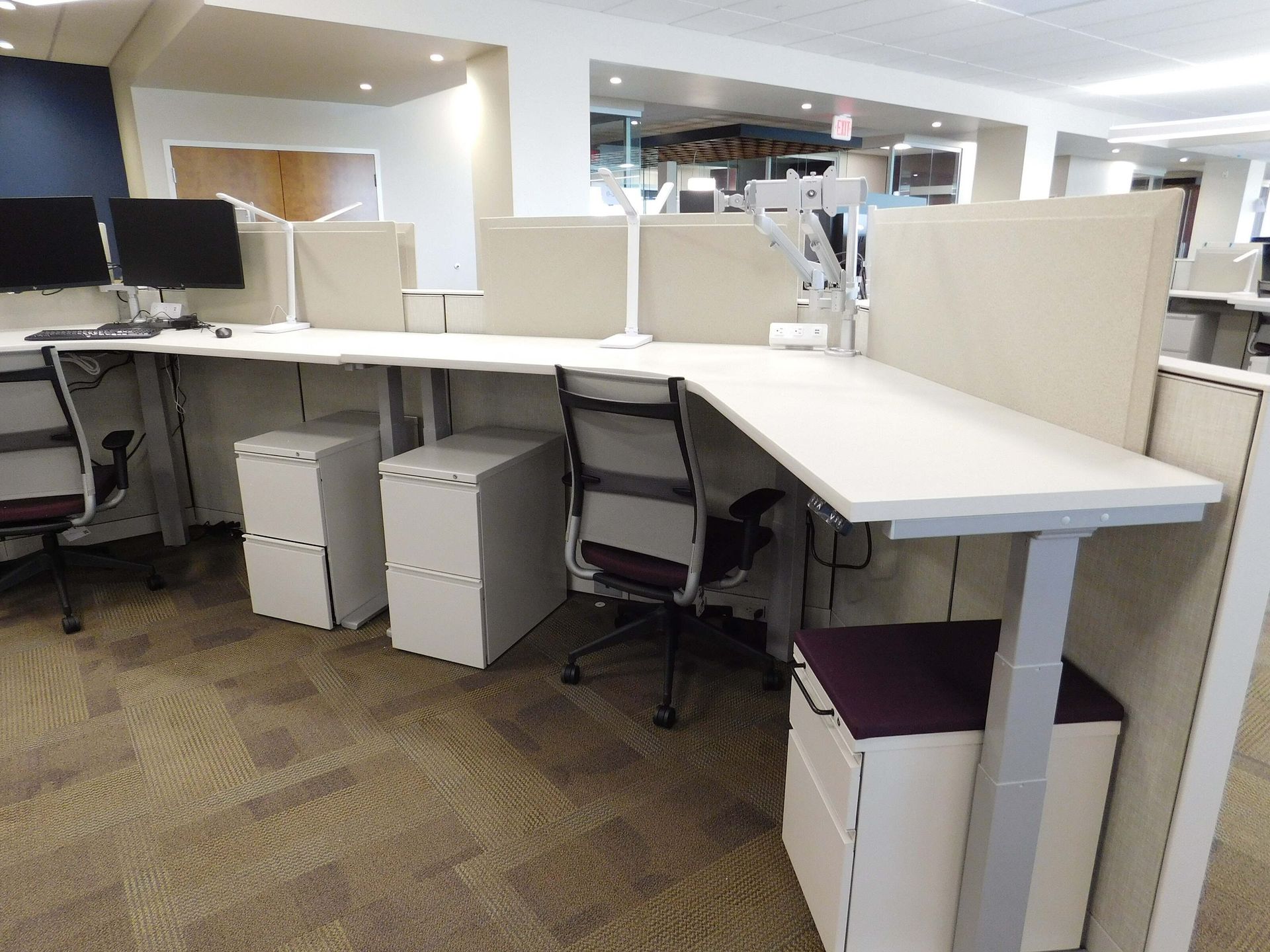 Open office cubicles with white desks, black chairs, and computer monitors under bright lighting.