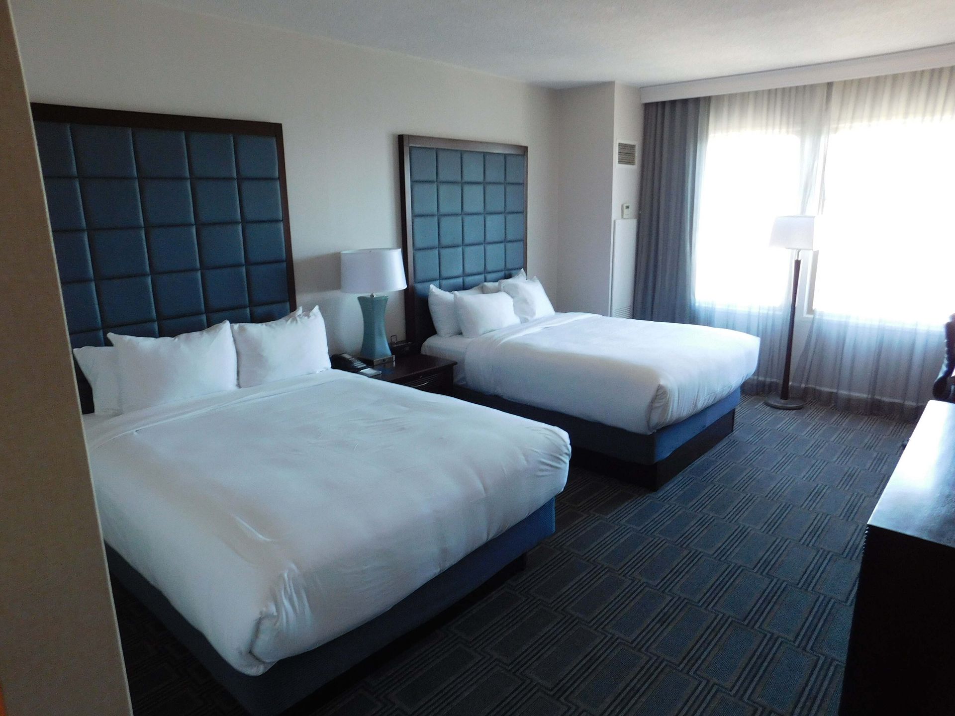 Hotel room with two neatly made white beds, dark headboards, and a bright window with curtains