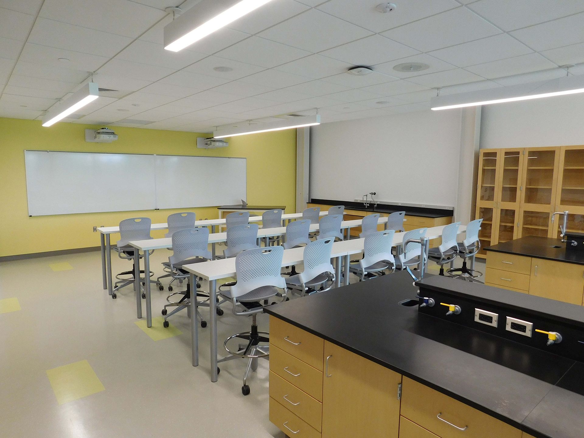 Bright classroom lab with desks, stools, whiteboard, cabinets, and a black worktable.