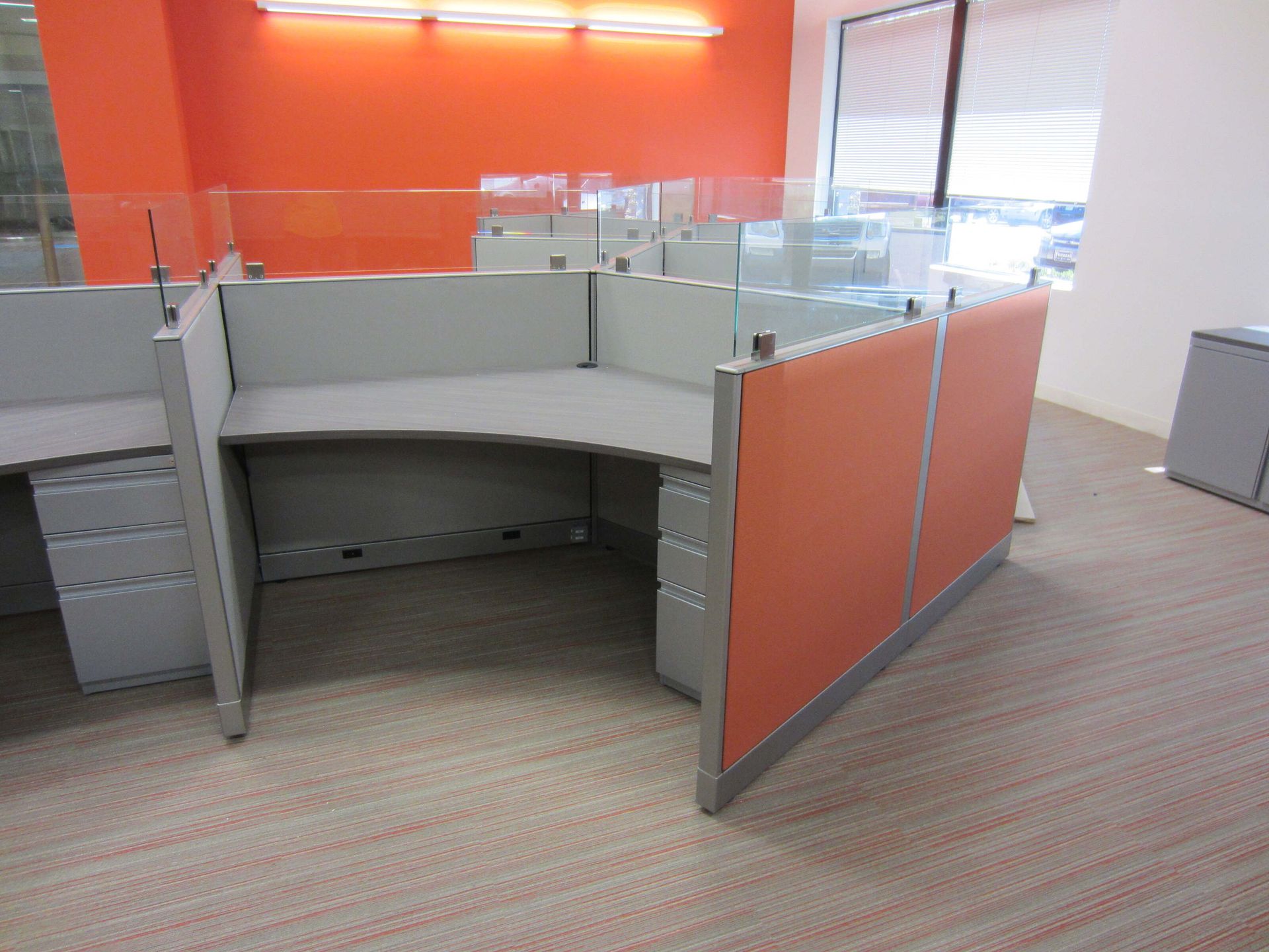 Empty office cubicle with orange and gray partitions, curved desk, and window blinds.