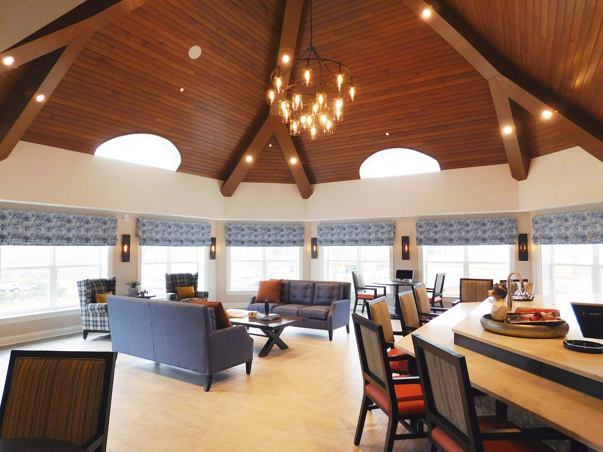 Spacious lounge with high wooden ceiling, chandelier, sofas, and large windows overlooking a bright patio