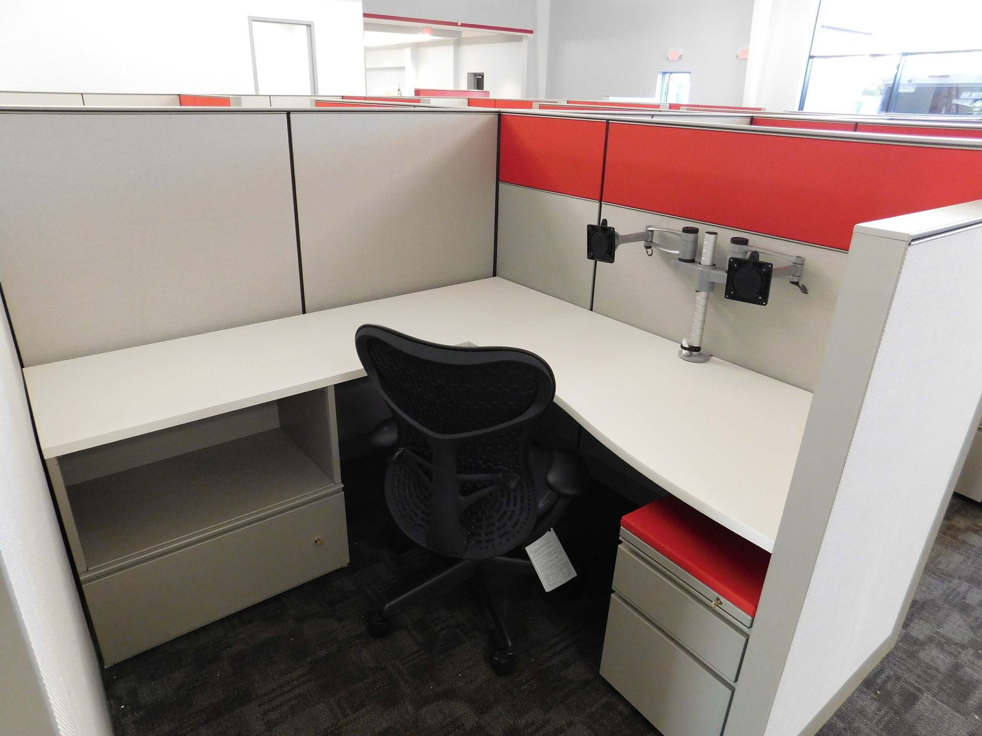 Empty office cubicle with black chair, white desks, and red trim.