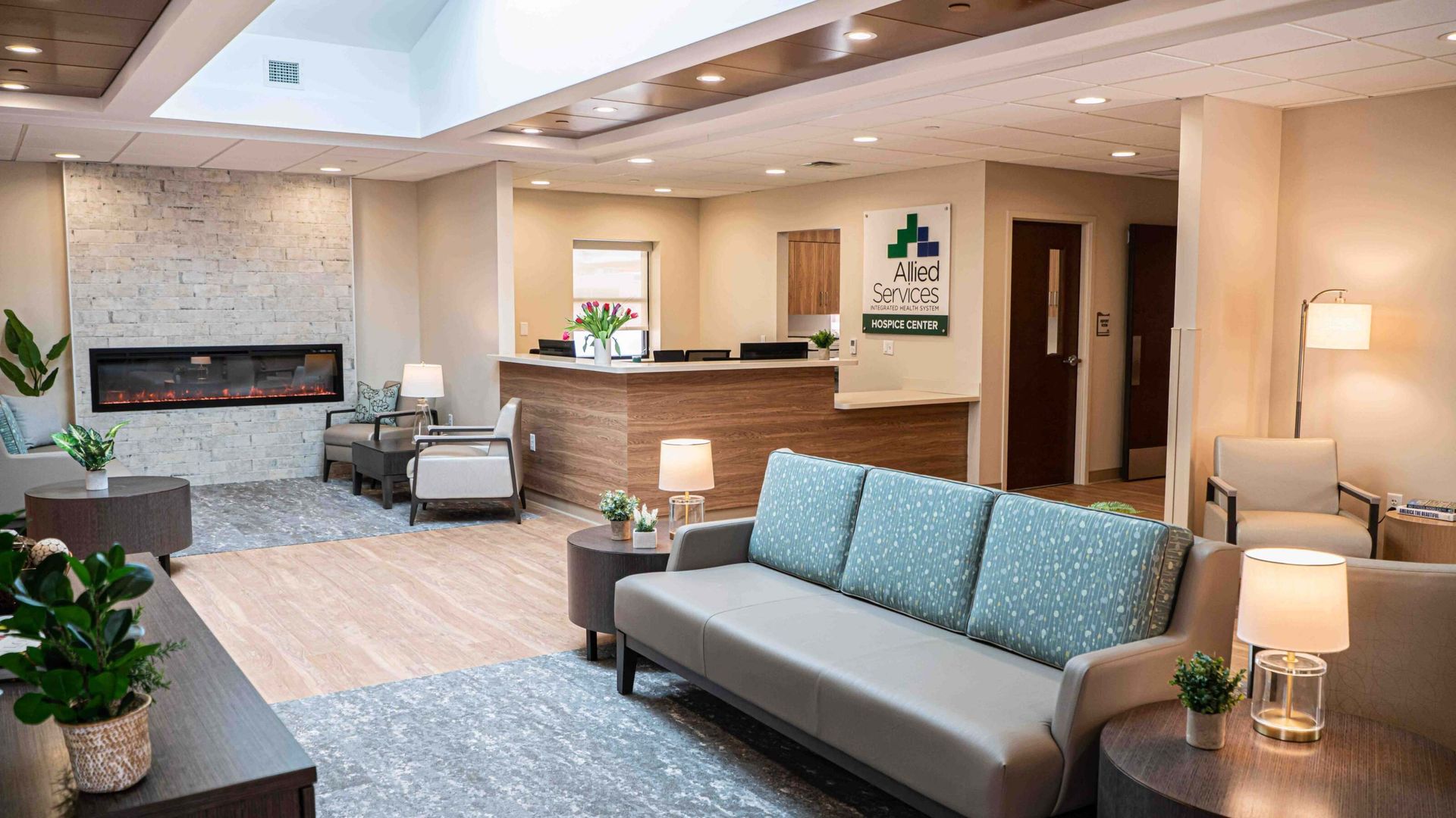 Modern hotel lobby with gray sofas, a fireplace, lamps, and a reception desk in warm neutral tones