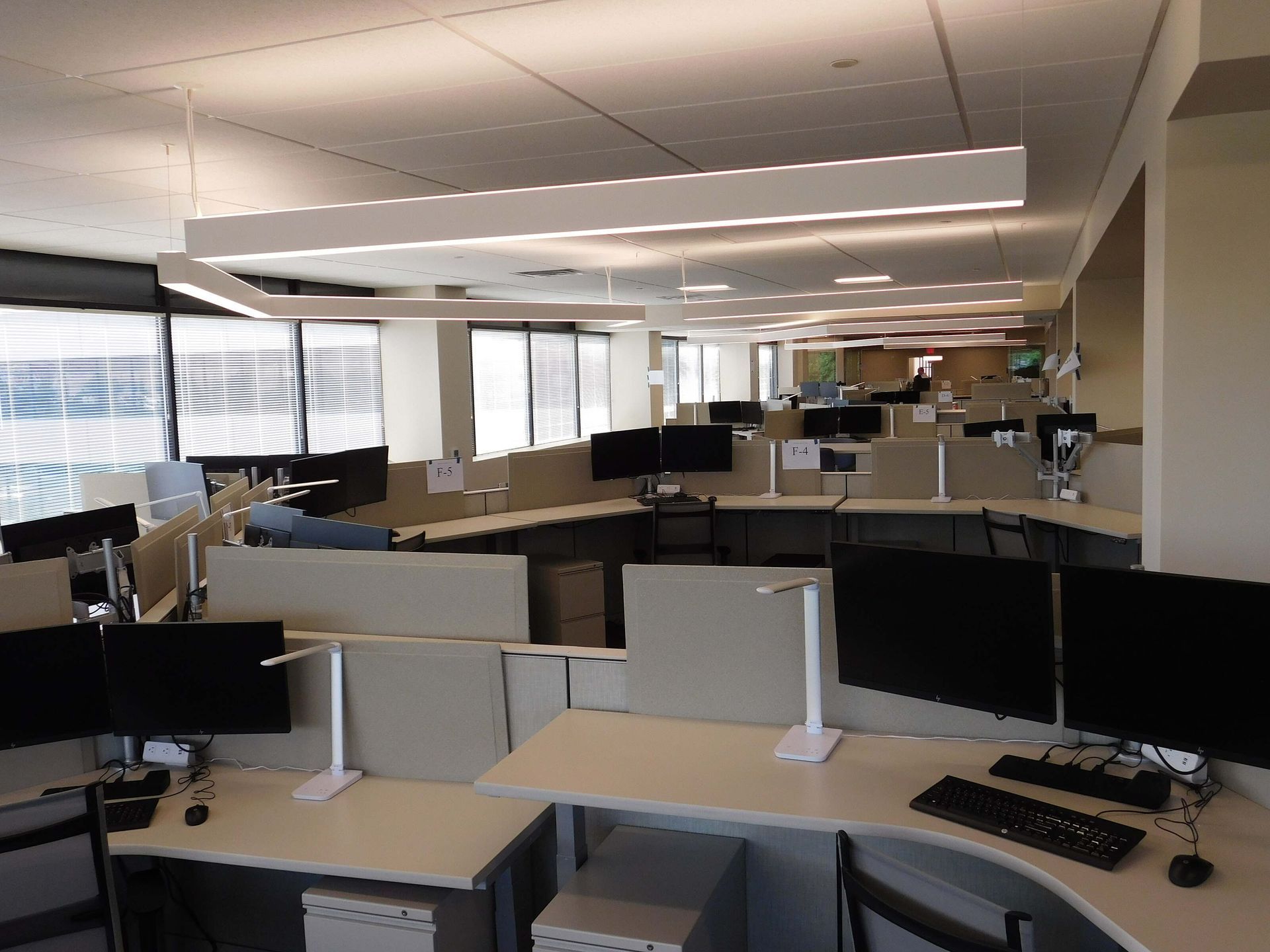 Empty office cubicles with desks, computers, and overhead lights in a modern open-plan workspace.