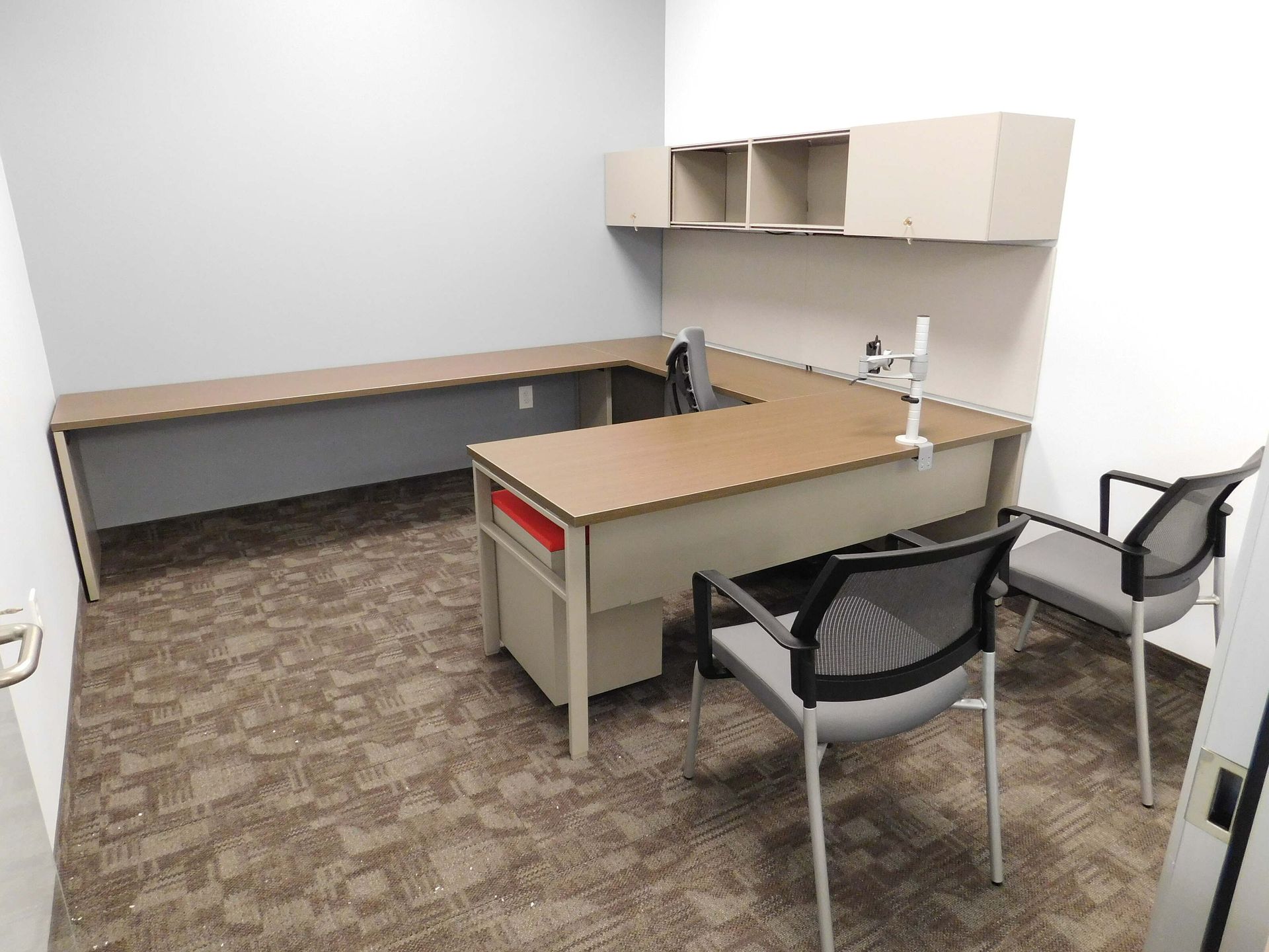 Empty office cubicle with beige desk, wall cabinets, and two black chairs on patterned carpet