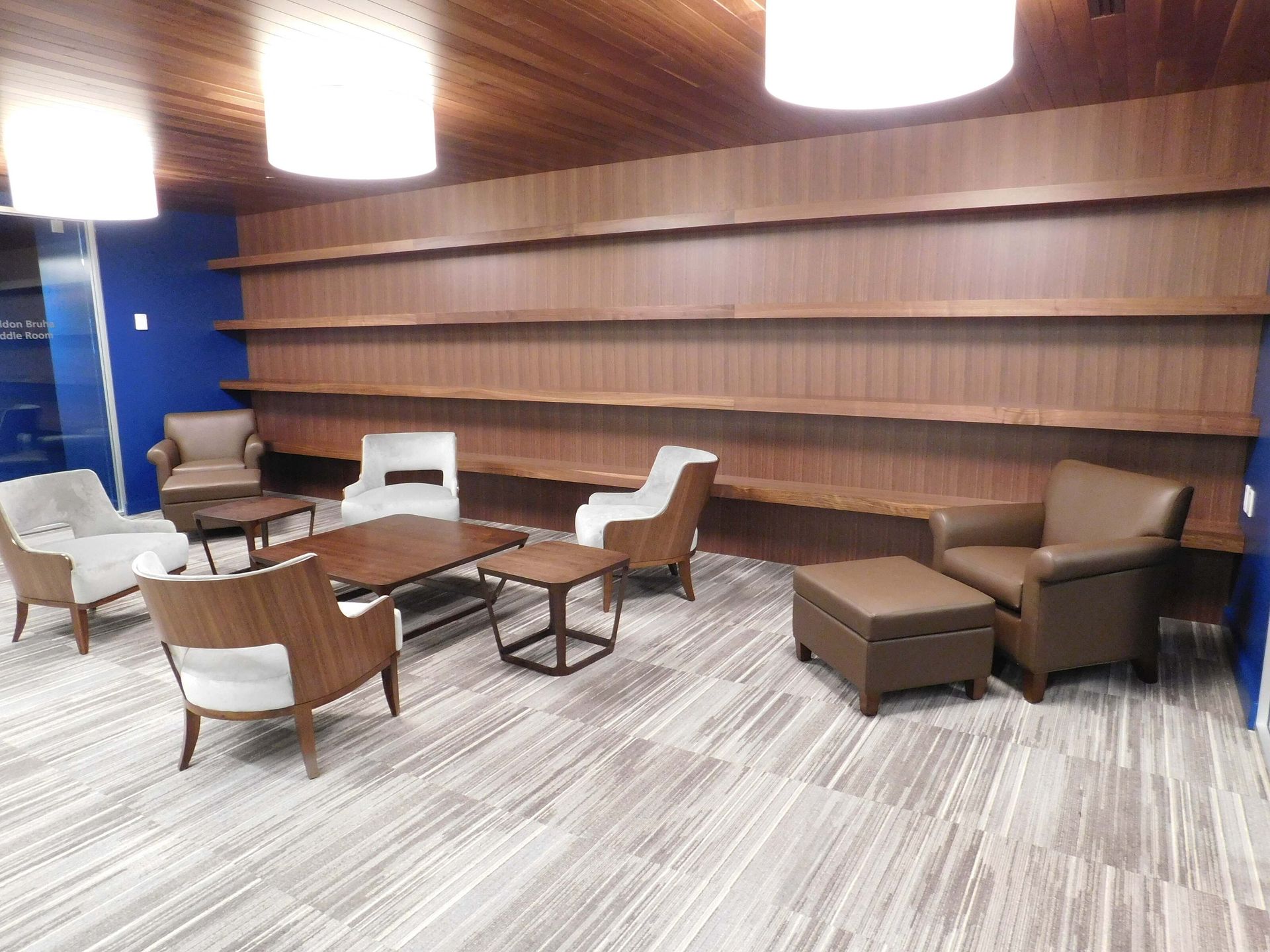 Modern lounge area with white and brown chairs, low tables, and a patterned carpet against wood-paneled walls