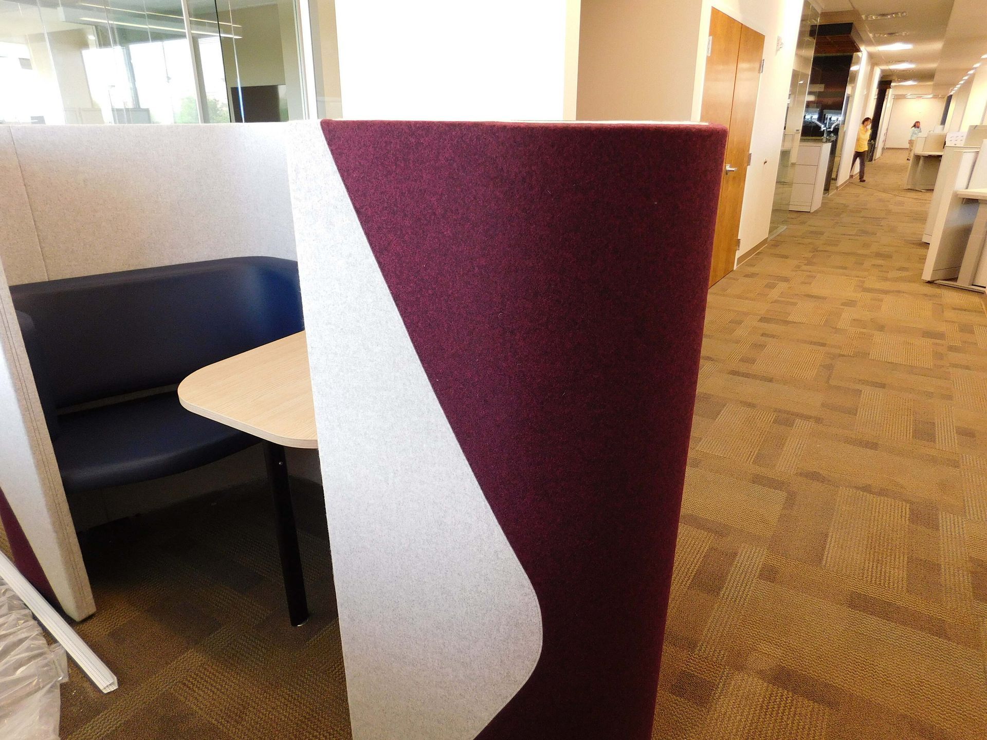 Office cubicle divider with burgundy and white panels beside a small table and booth seating in a hallway