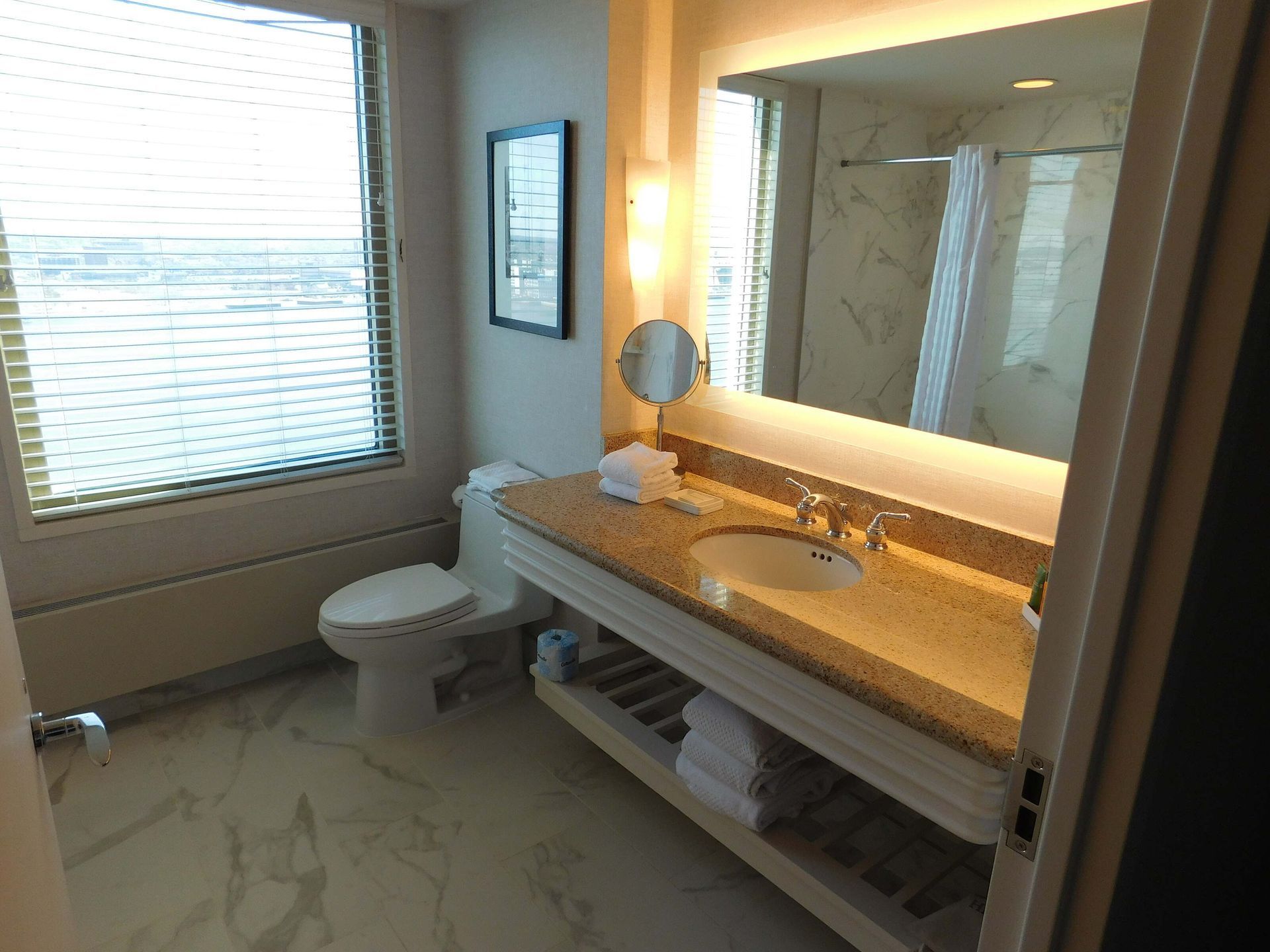 Bright bathroom with window, toilet, and marble vanity with lit mirror above sink.