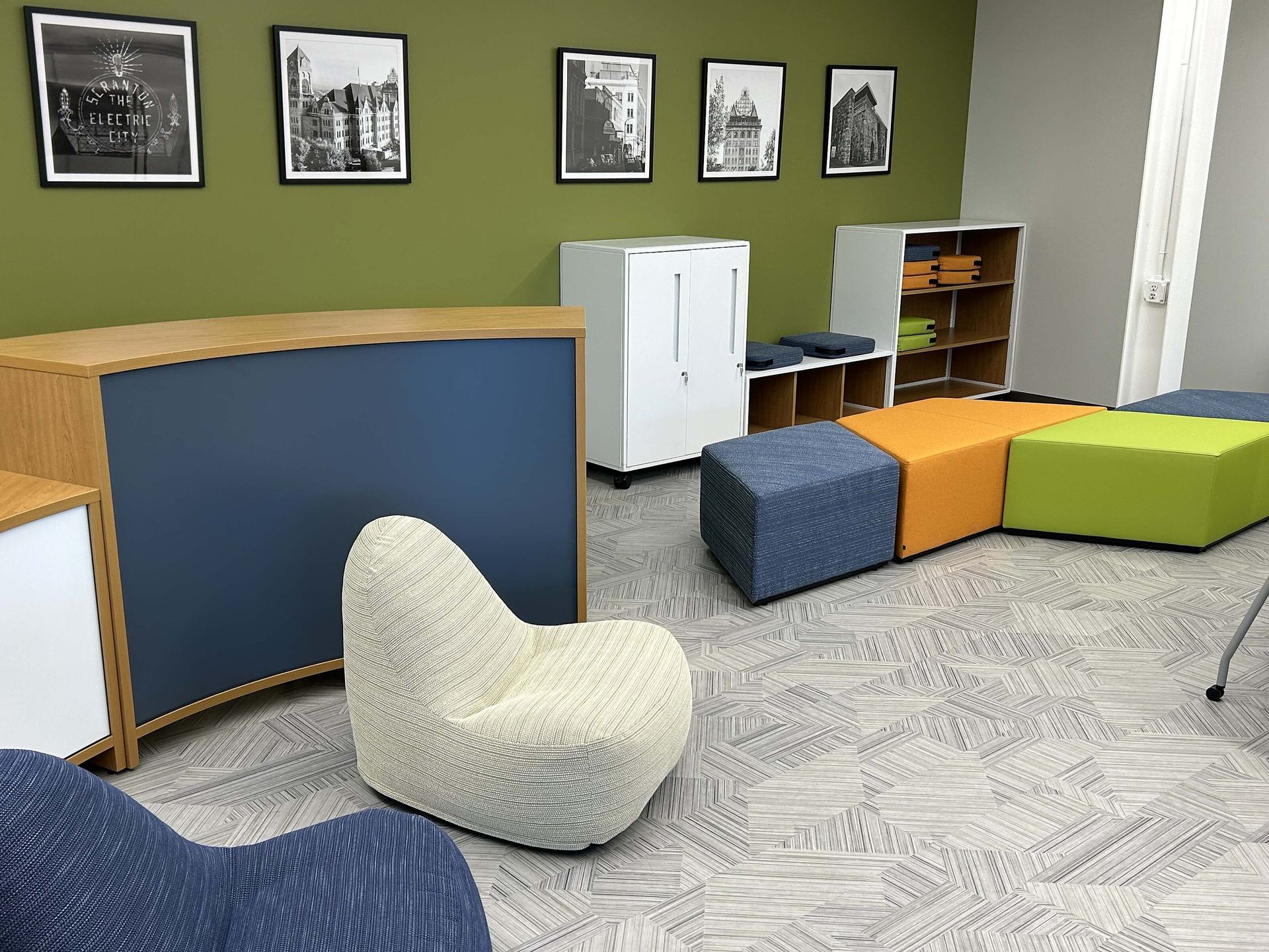 Modern classroom corner with chairs, cabinets, wall photos, and colorful seating on gray carpet