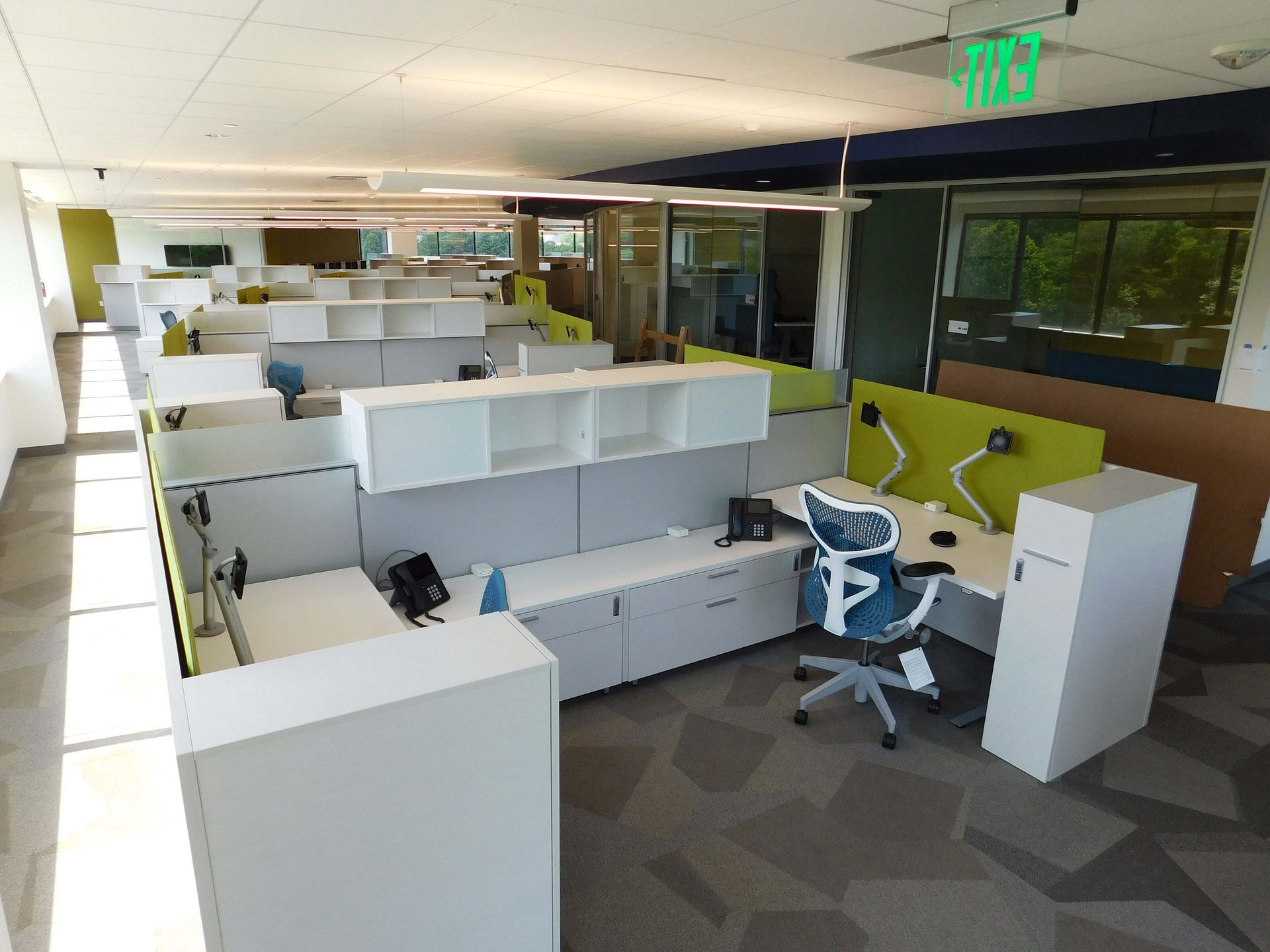 Empty modern office cubicles with white desks, gray carpet, and a lime-green chair near windows.