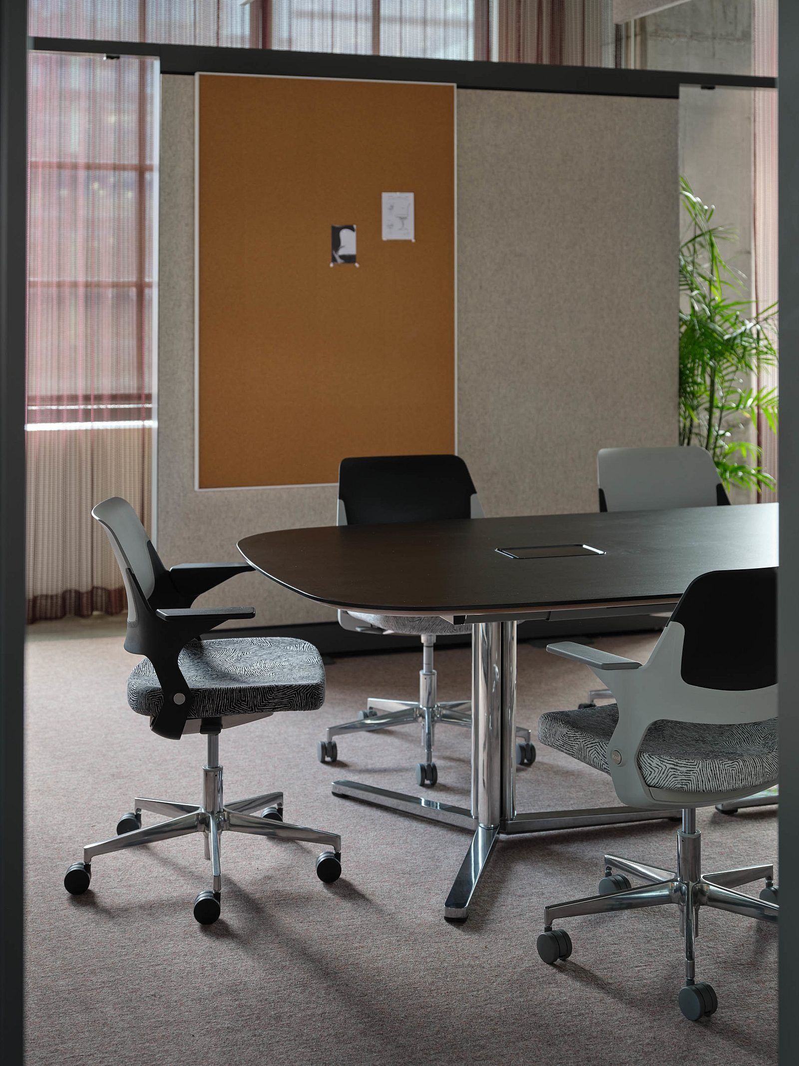 Modern conference room with oval table, office chairs, corkboard, and large windows.
