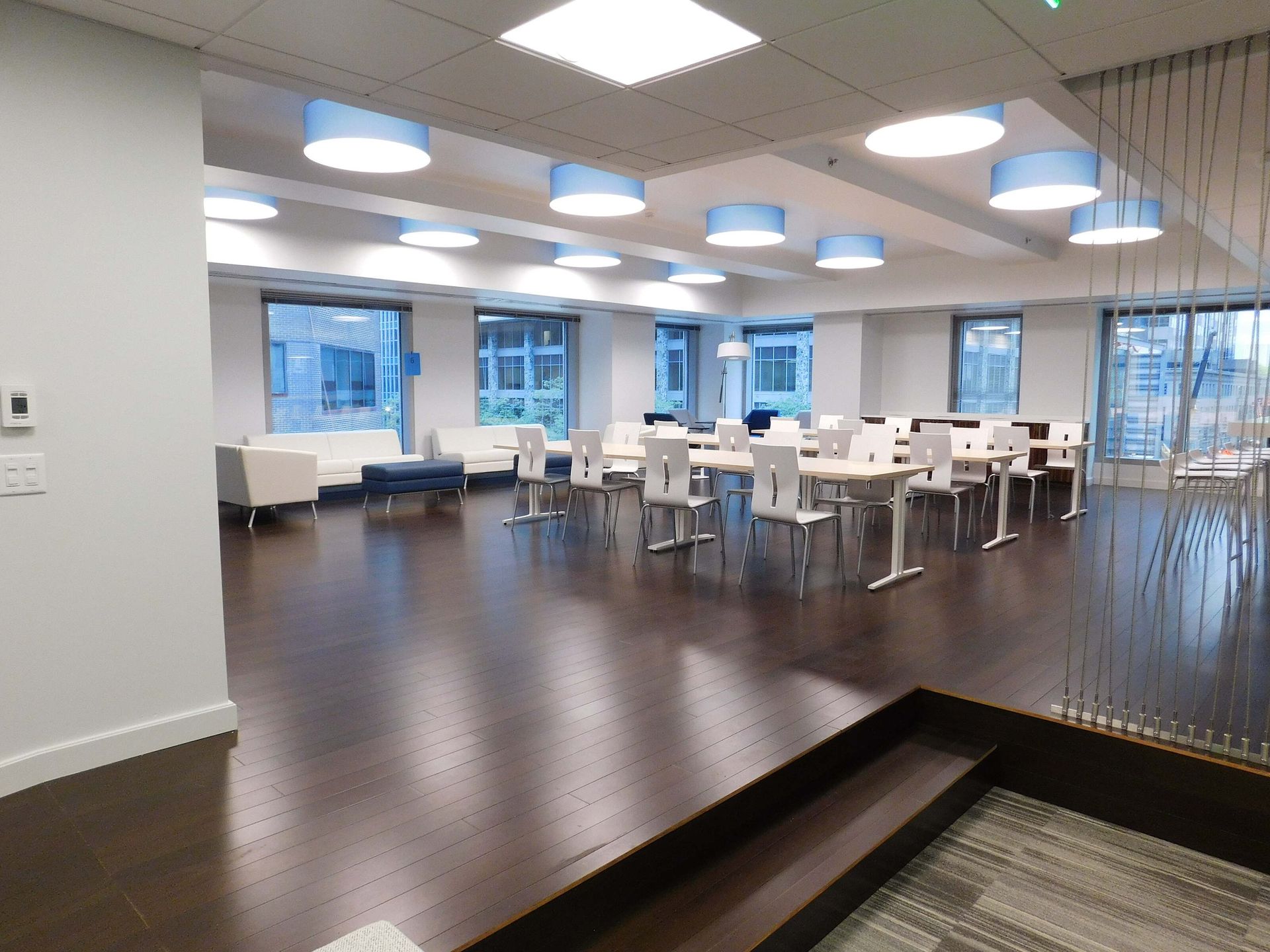 Modern open office with tables, chairs, dark wood floor, and large windows under round ceiling lights