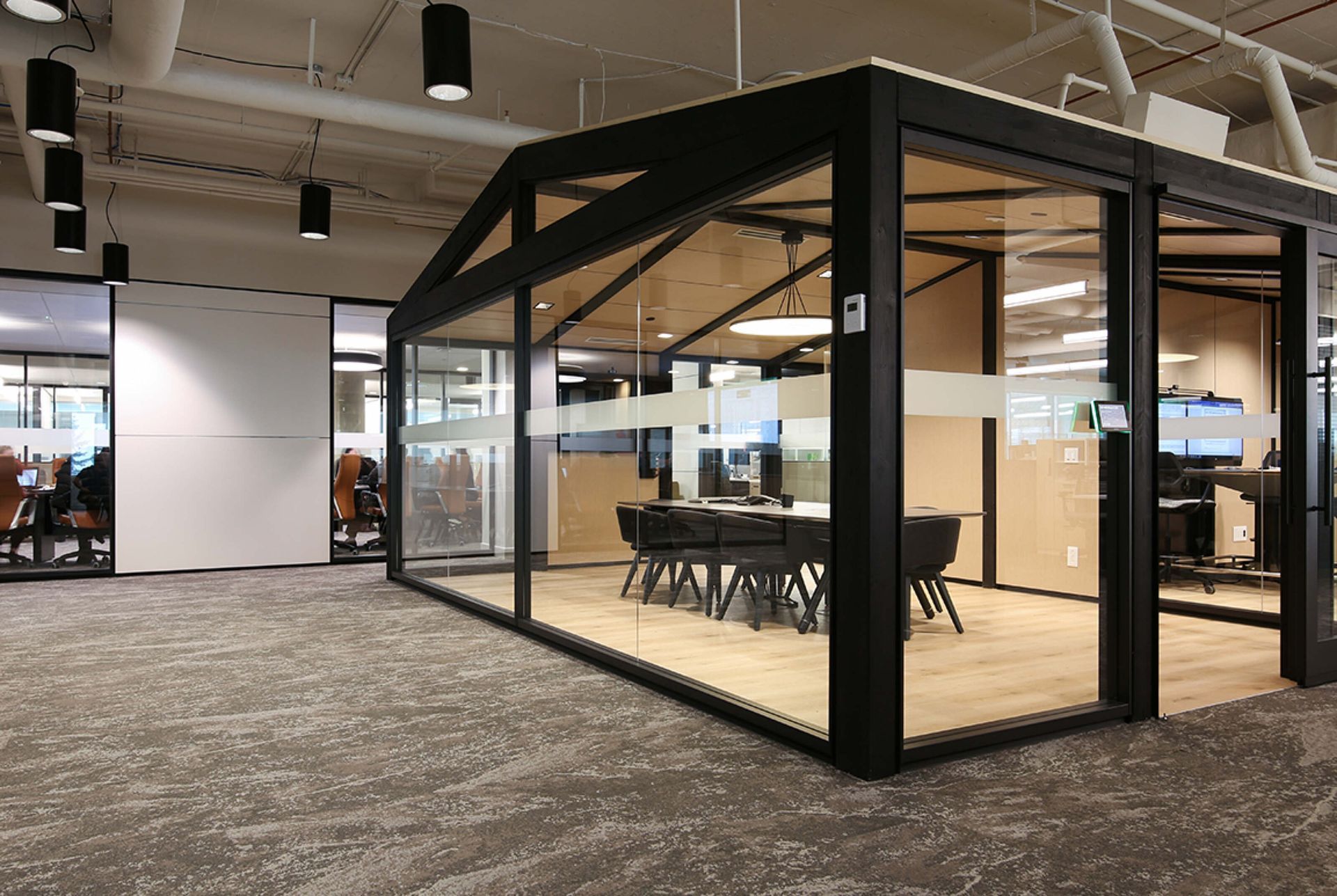 Glass-walled office meeting room with black frame inside a carpeted open workspace