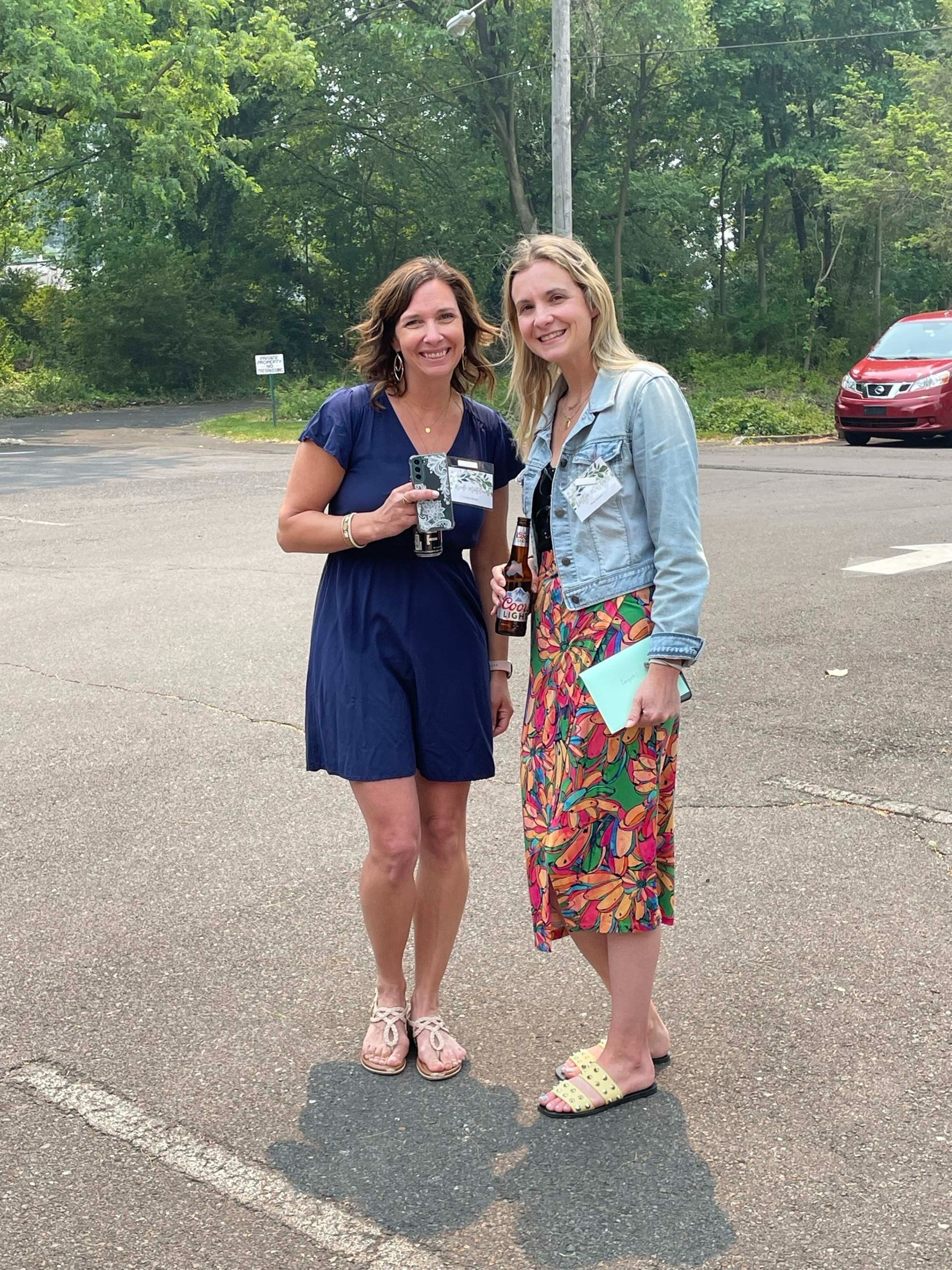 Two women standing together outdoors in a parking lot, smiling and holding drinks.