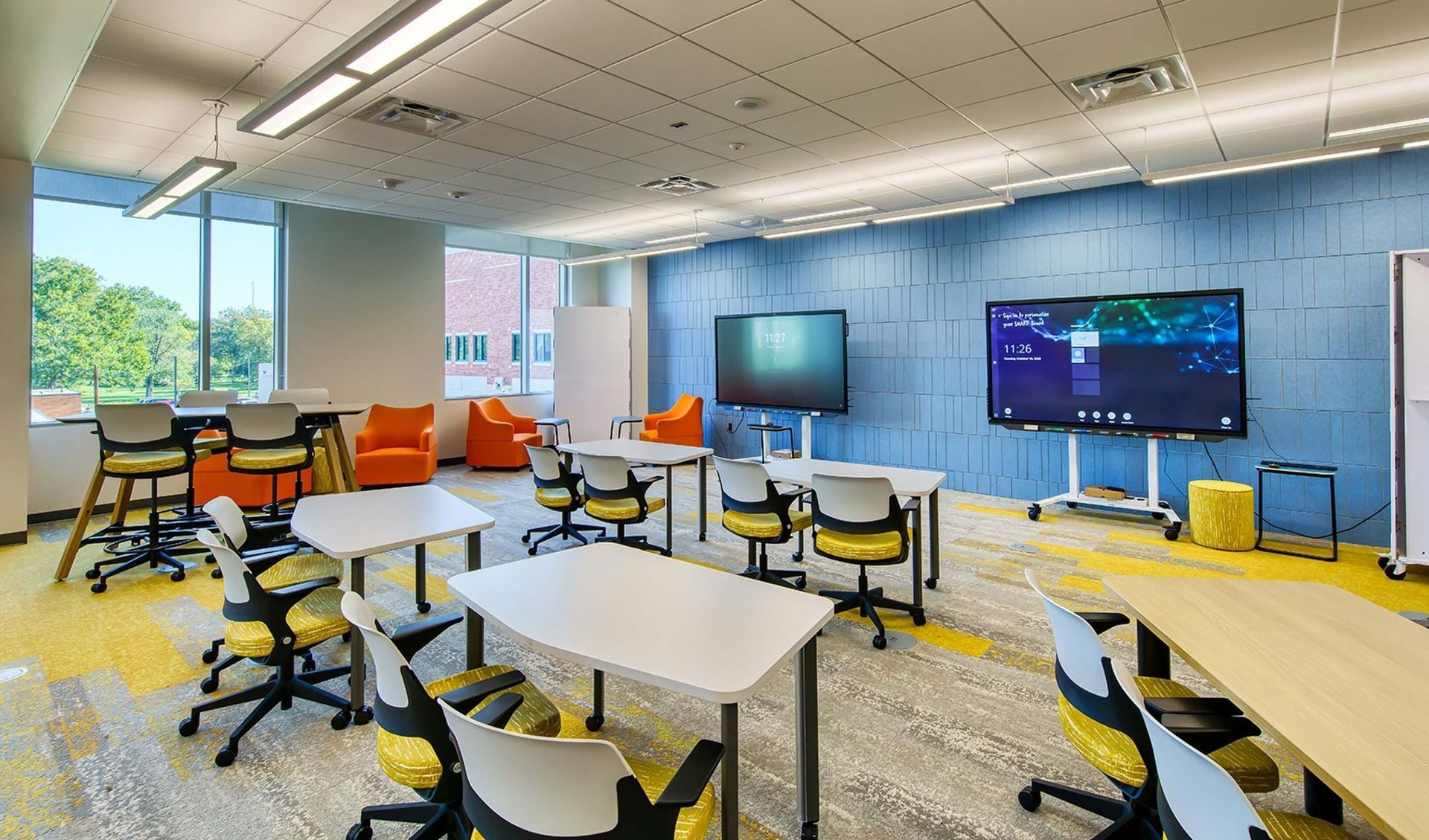 Modern classroom with tables, chairs, yellow rugs, and wall-mounted screens in a bright, open learning space