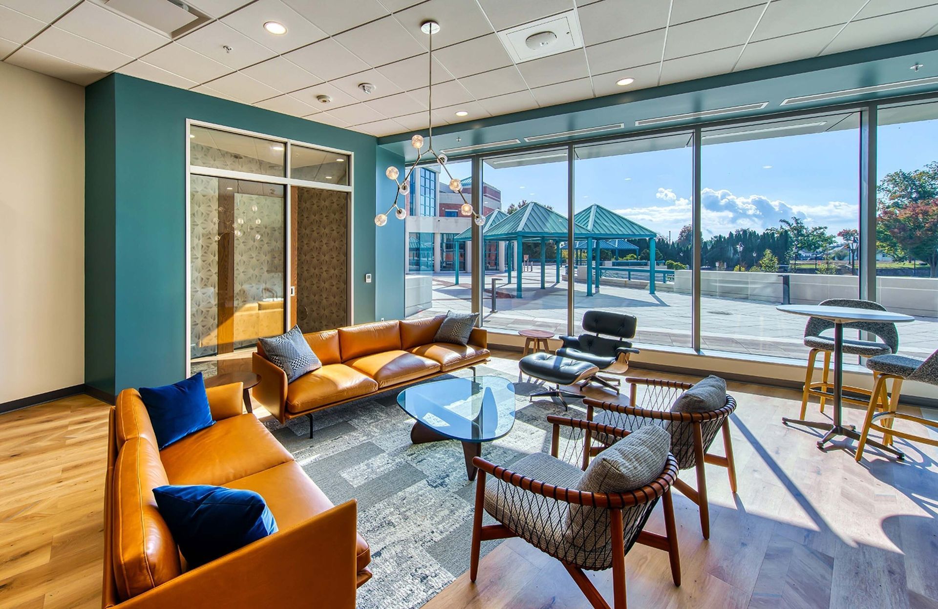 Bright modern lounge with tan sofas, tables, and large windows overlooking a sunny outdoor patio and water.