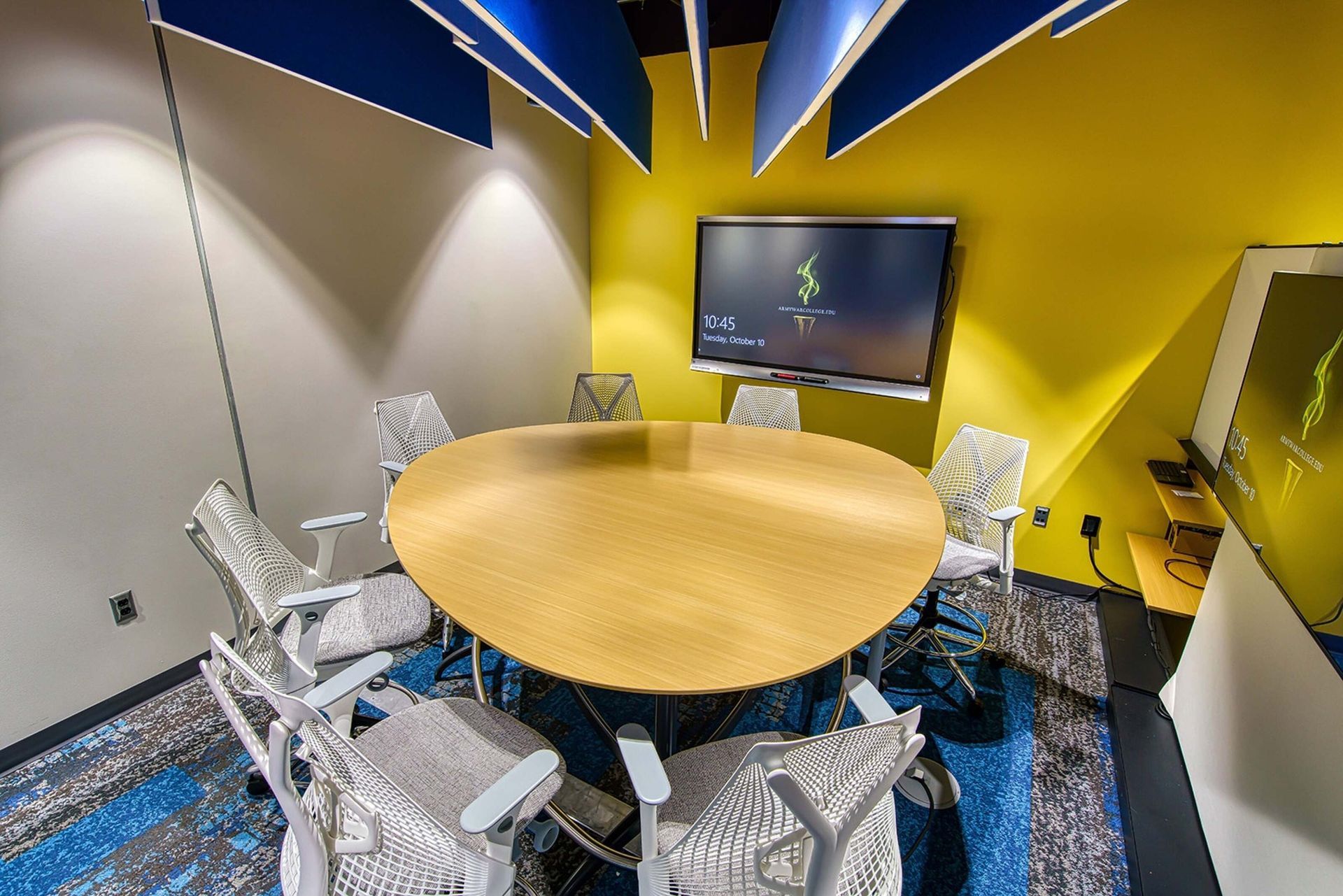 Modern conference room with round wooden table, white chairs, blue-and-yellow ceiling, and wall-mounted screen
