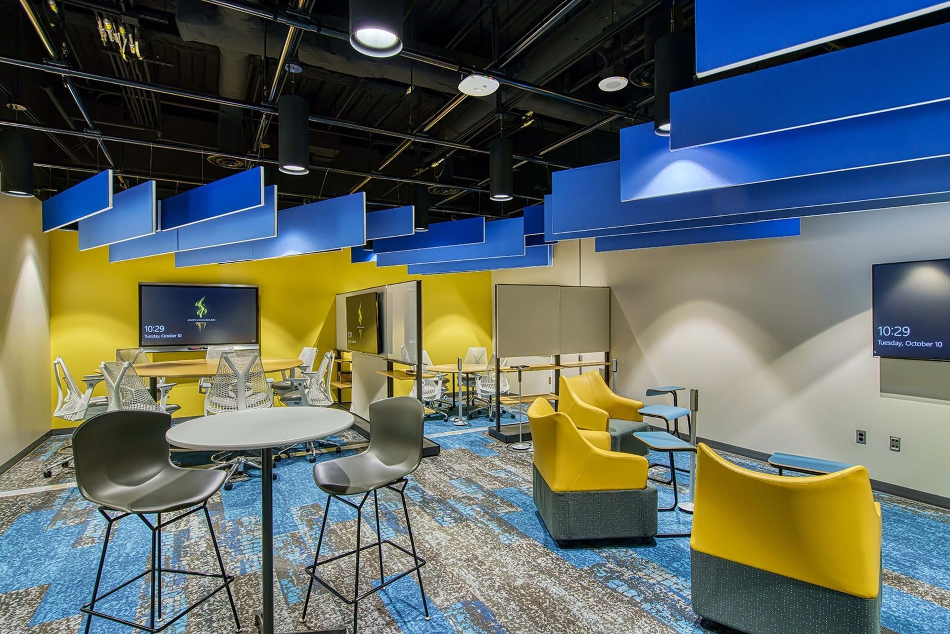 Modern lounge with yellow and blue seating, round tables, and a patterned carpet under hanging blue panels
