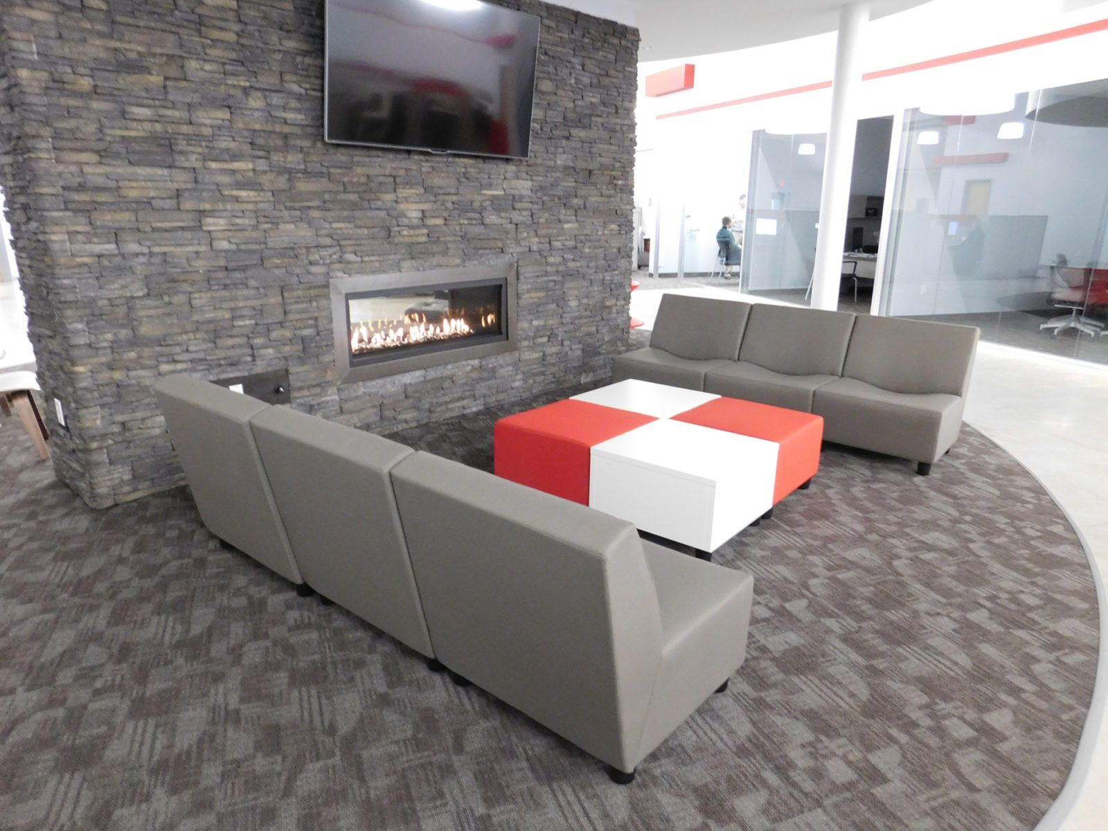 Modern lounge with gray sofas around a red-and-white coffee table, stone fireplace, and TV monitor.