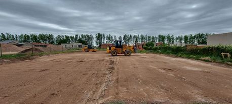 Una excavadora nivela un amplio terreno de construcción bajo un cielo nublado, con árboles al fondo.
