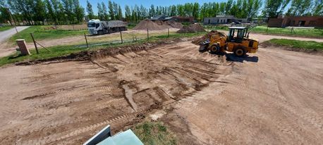 Una cargadora amarilla nivela un terreno de tierra con árboles y vehículos estacionados al fondo.
