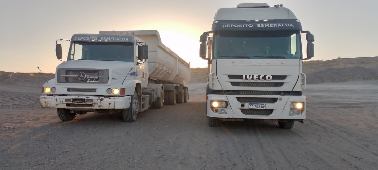 Dos camiones volquete Volvo blancos estacionados uno al lado del otro en un camino de tierra al atardecer.