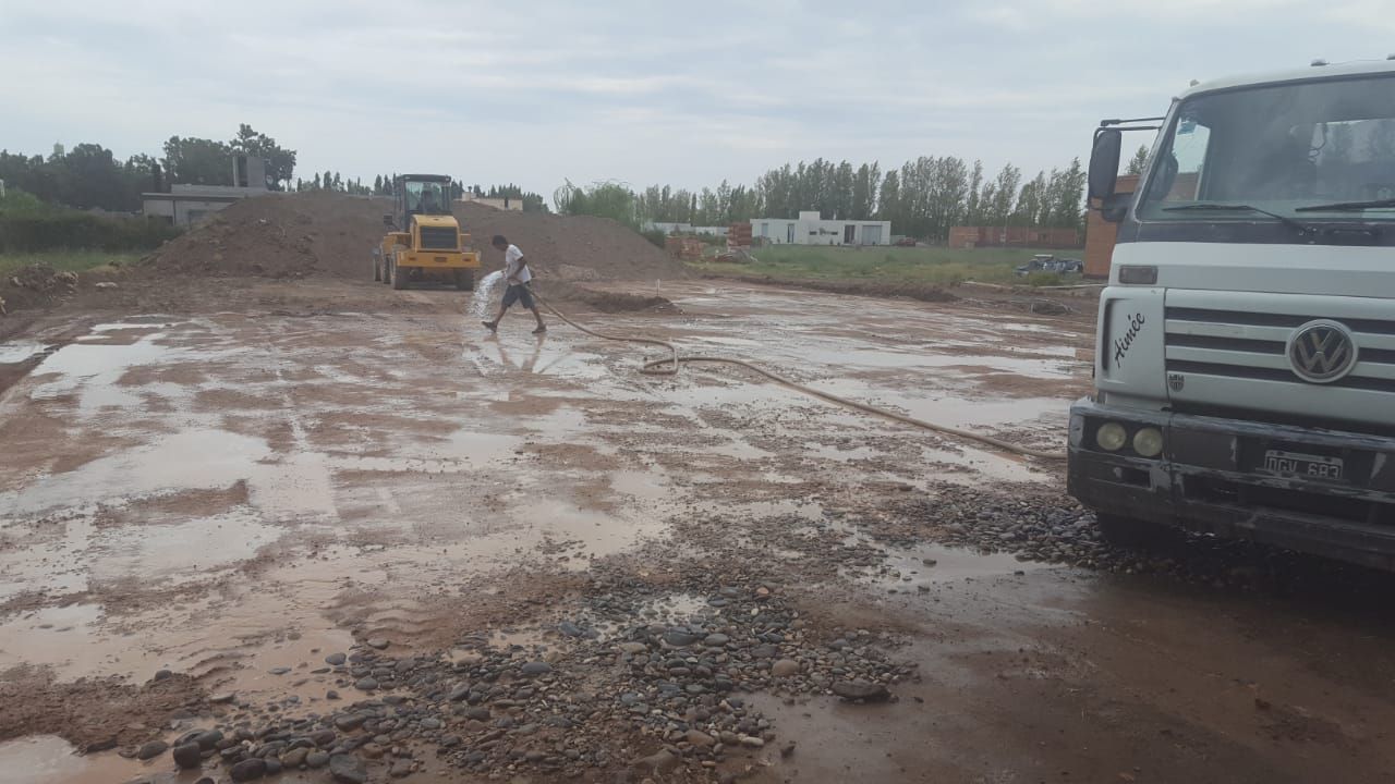 Obra en construcción embarrada con un trabajador, una excavadora amarilla y un camión a la derecha.