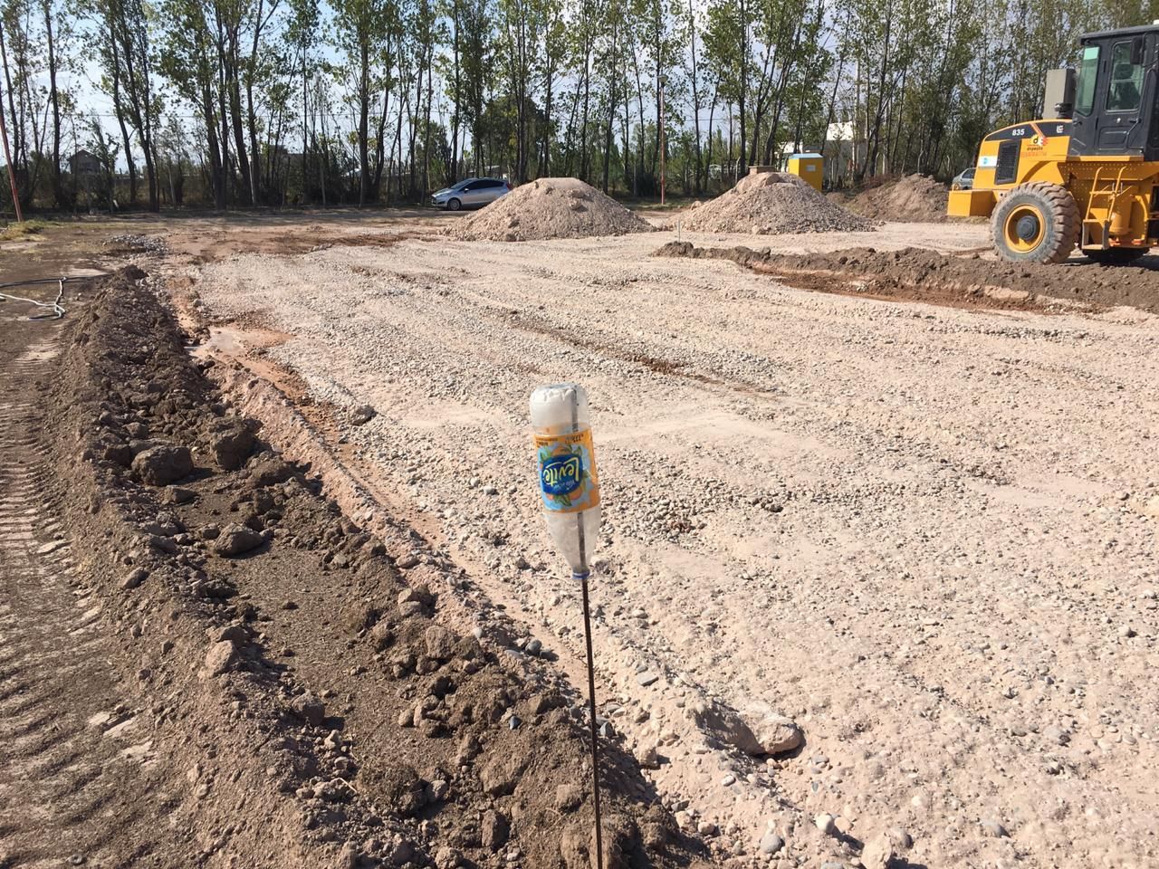Obra en construcción de un camino de tierra con grava, una estaca de topografía, montones de tierra y maquinaria pesada.