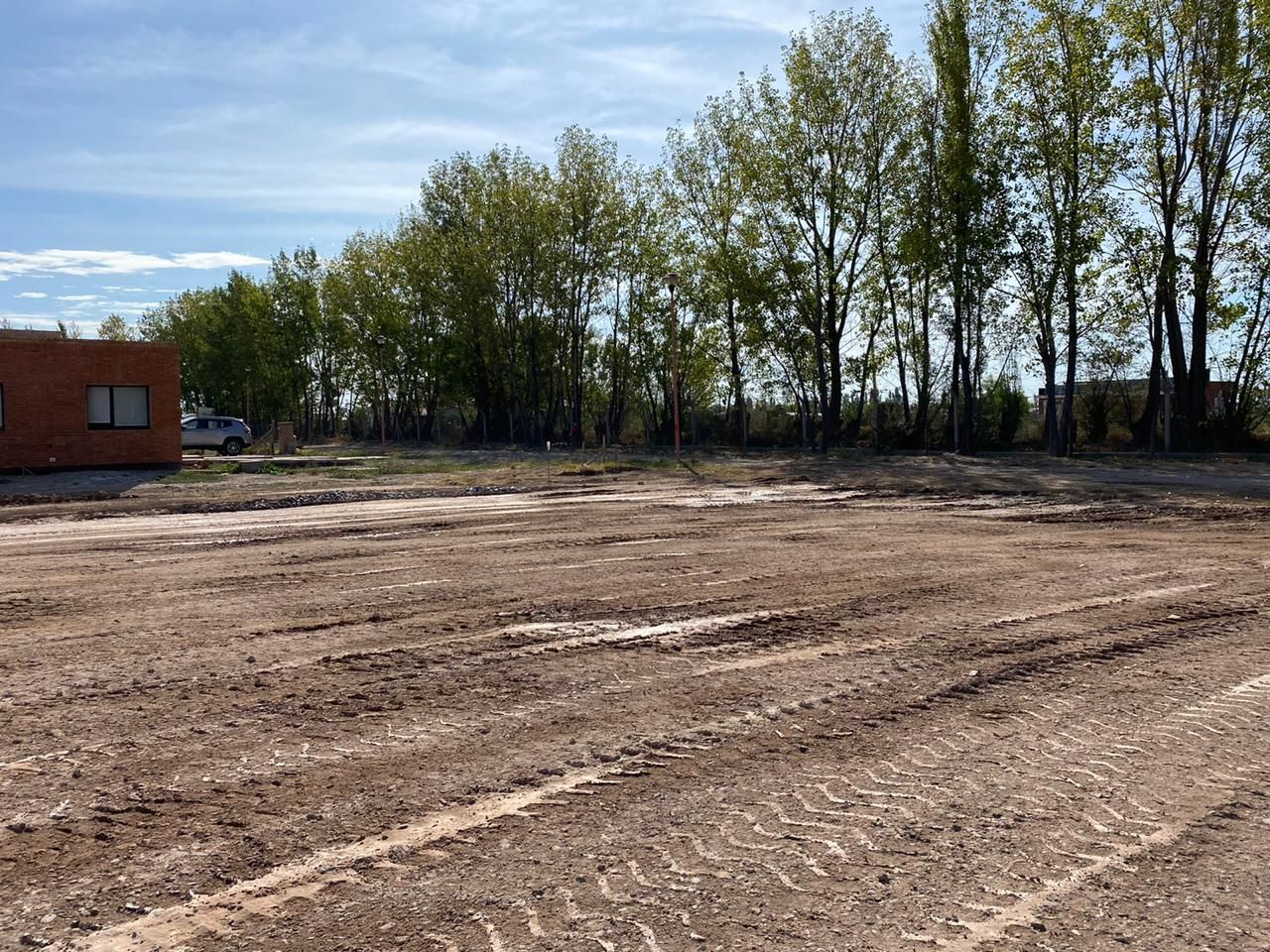 Terreno despejado con huellas de neumáticos, árboles al fondo y un pequeño edificio de ladrillos a la izquierda.