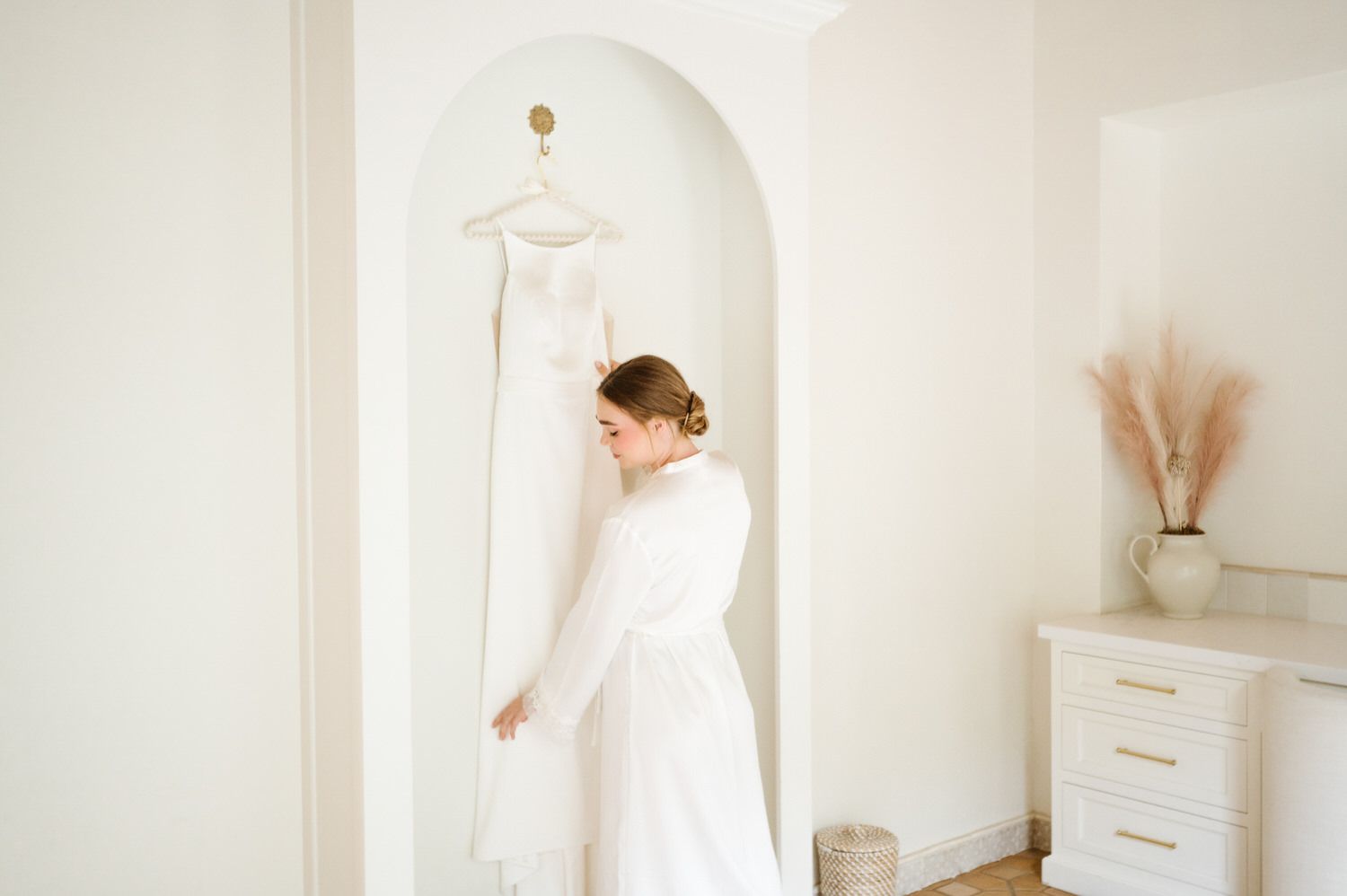 A woman in a white dress is looking at her wedding dress hanging on a wall.