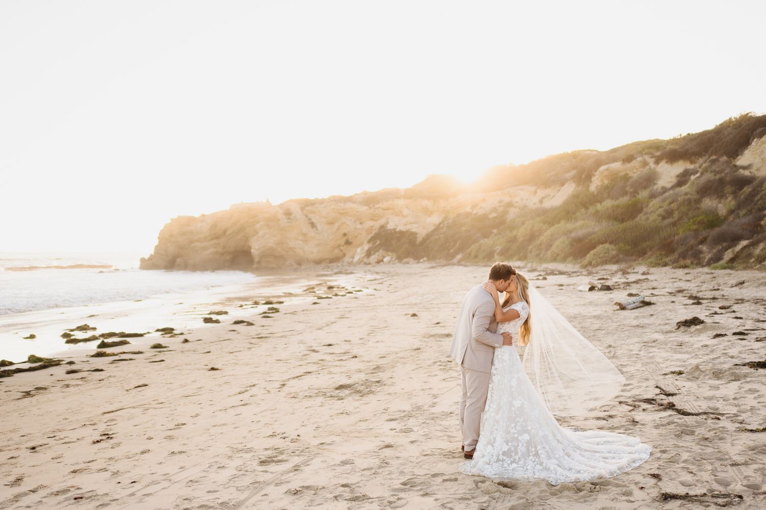 A bride and groom are kissing on the beach at sunset.