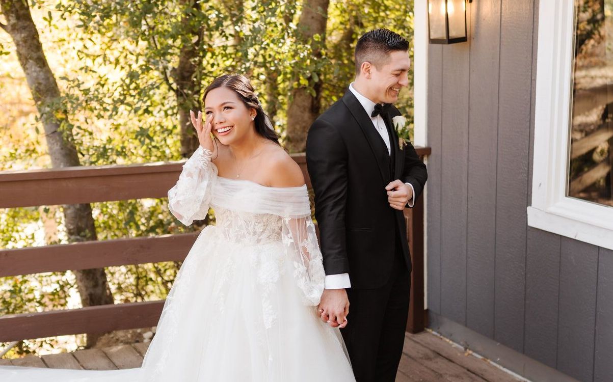A bride and groom are standing next to each other on a balcony holding hands crying and laughing.
