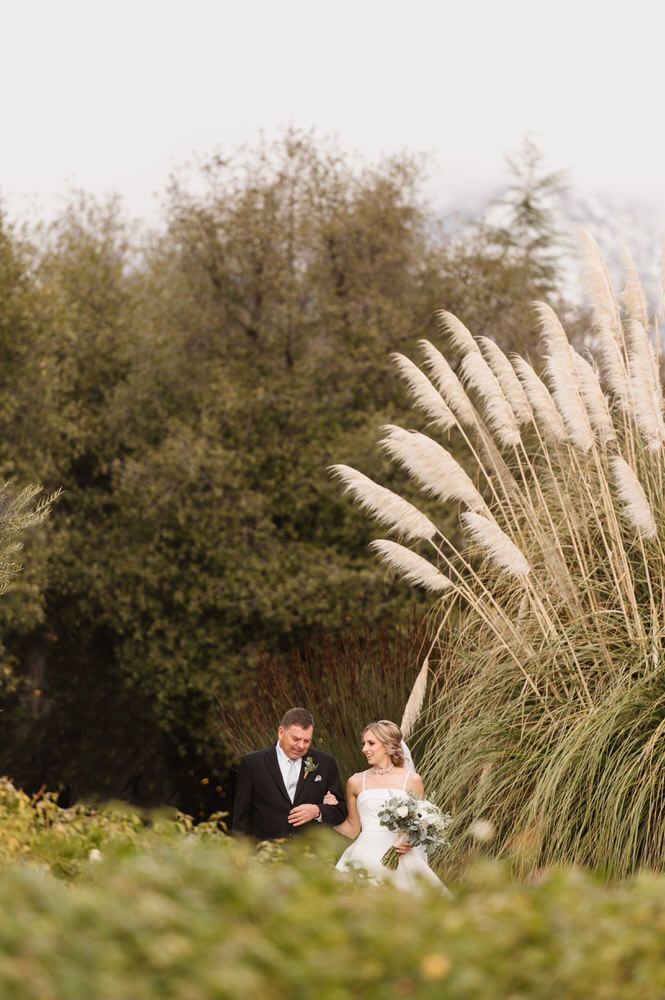 Bride and father standing at wedding venue, Serendipity Gardens with tall grasses and trees, holding flowers outdoors