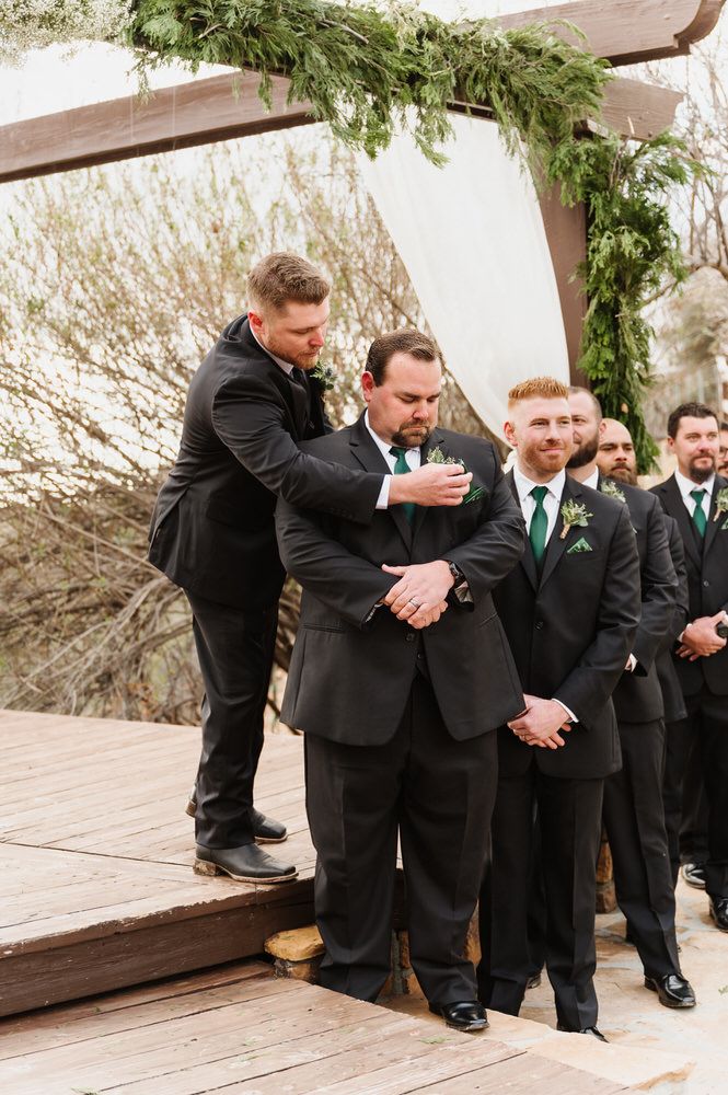 Groomsmen in black suits standing on a wooden platform during a wedding ceremony outdoors