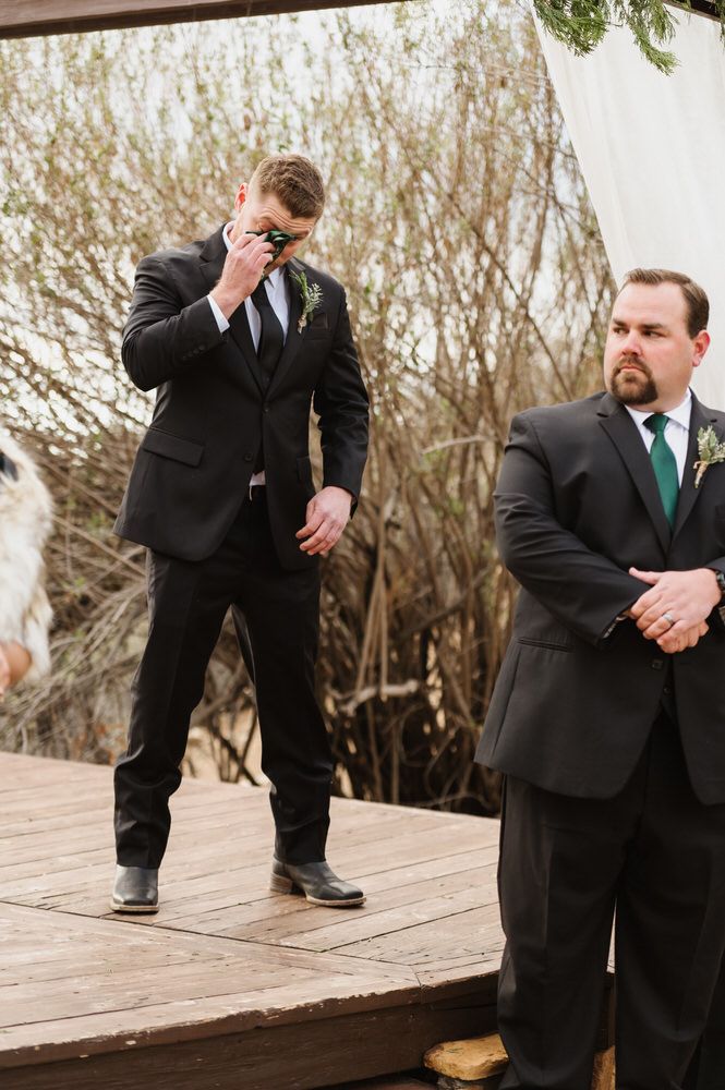 Two men in suits stand on a wooden deck outdoors, one adjusting his tie, with tall grasses behind them.