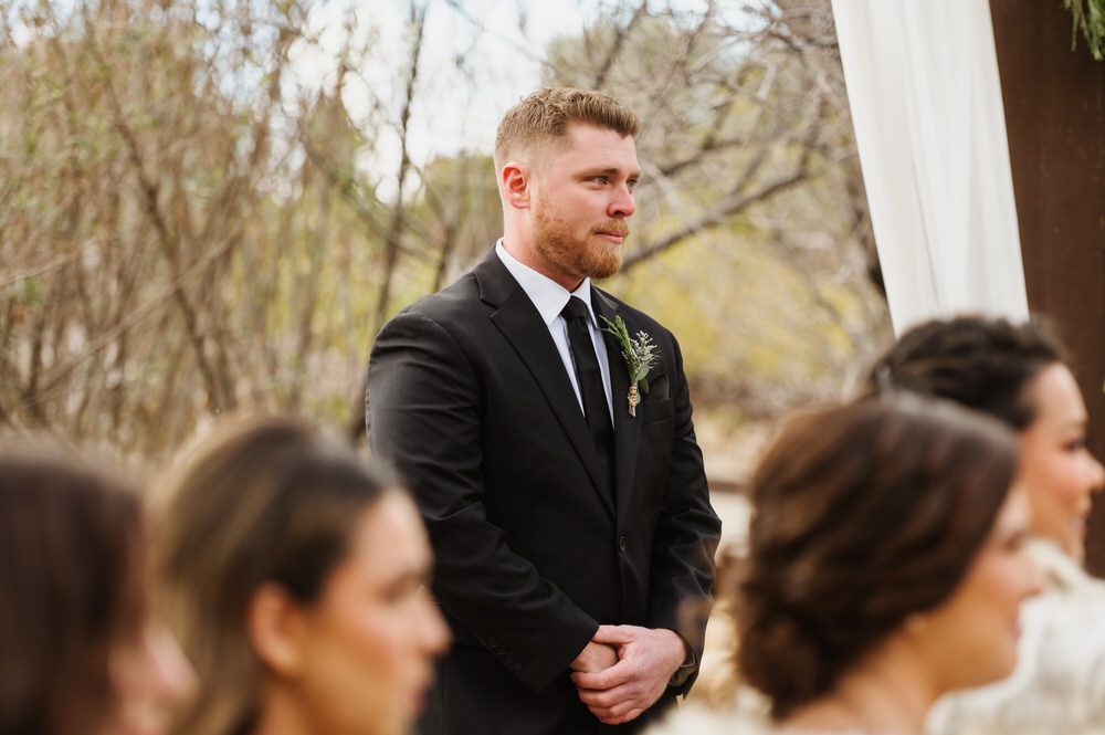 Groom in black suit stands at outdoor wedding ceremony, with guests blurred in foreground.