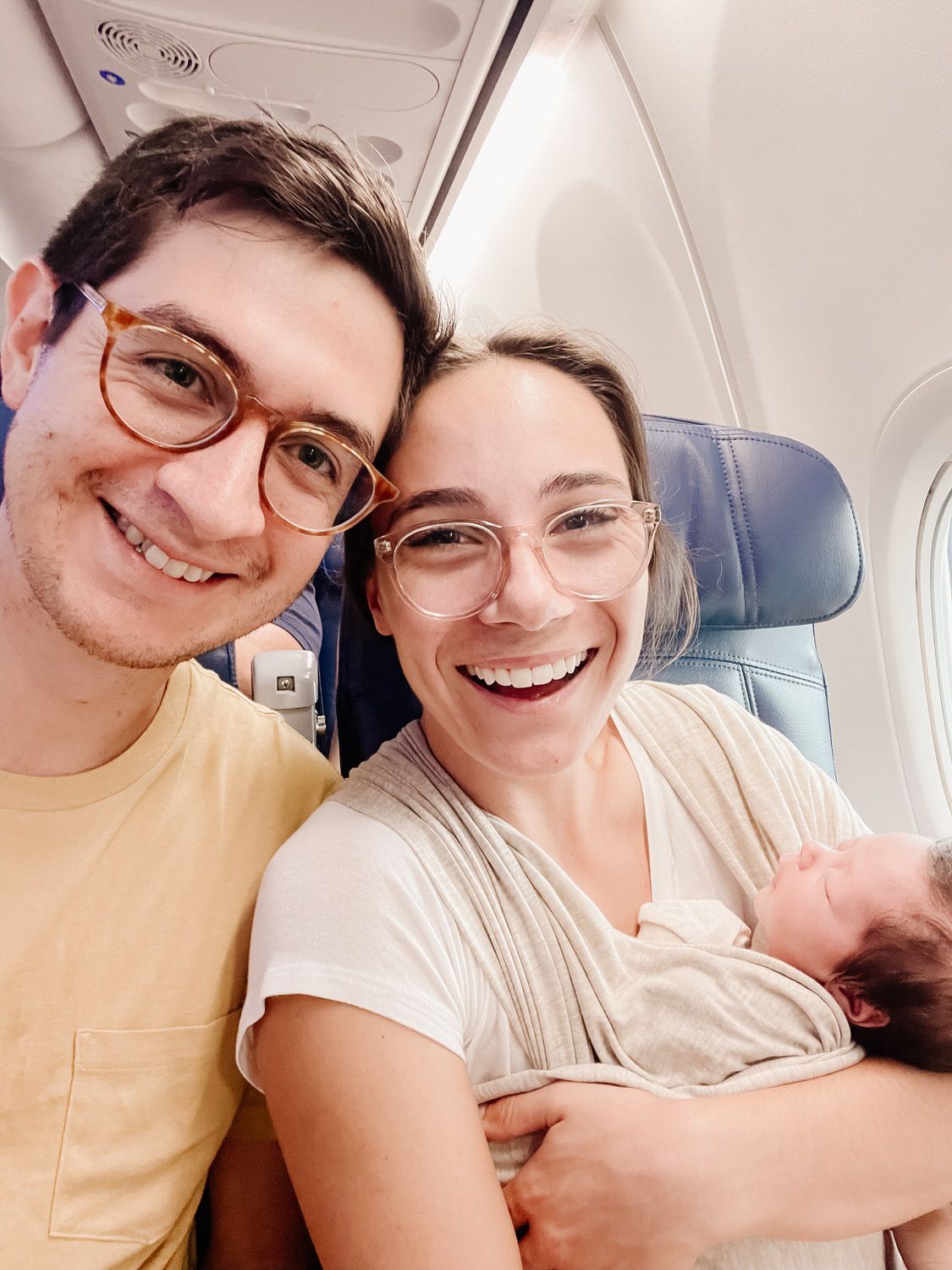 Smiling couple taking a selfie on an airplane, holding a newborn in a seat