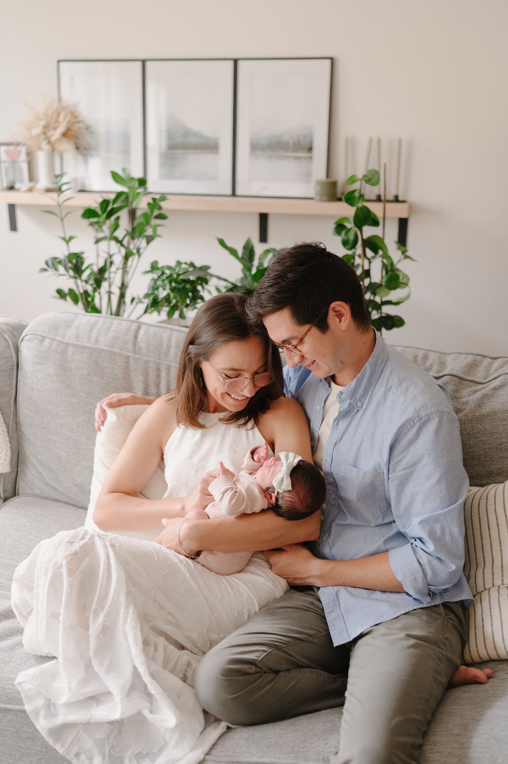 Couple sitting on a couch, gently holding and looking at a sleeping baby in a bright living room