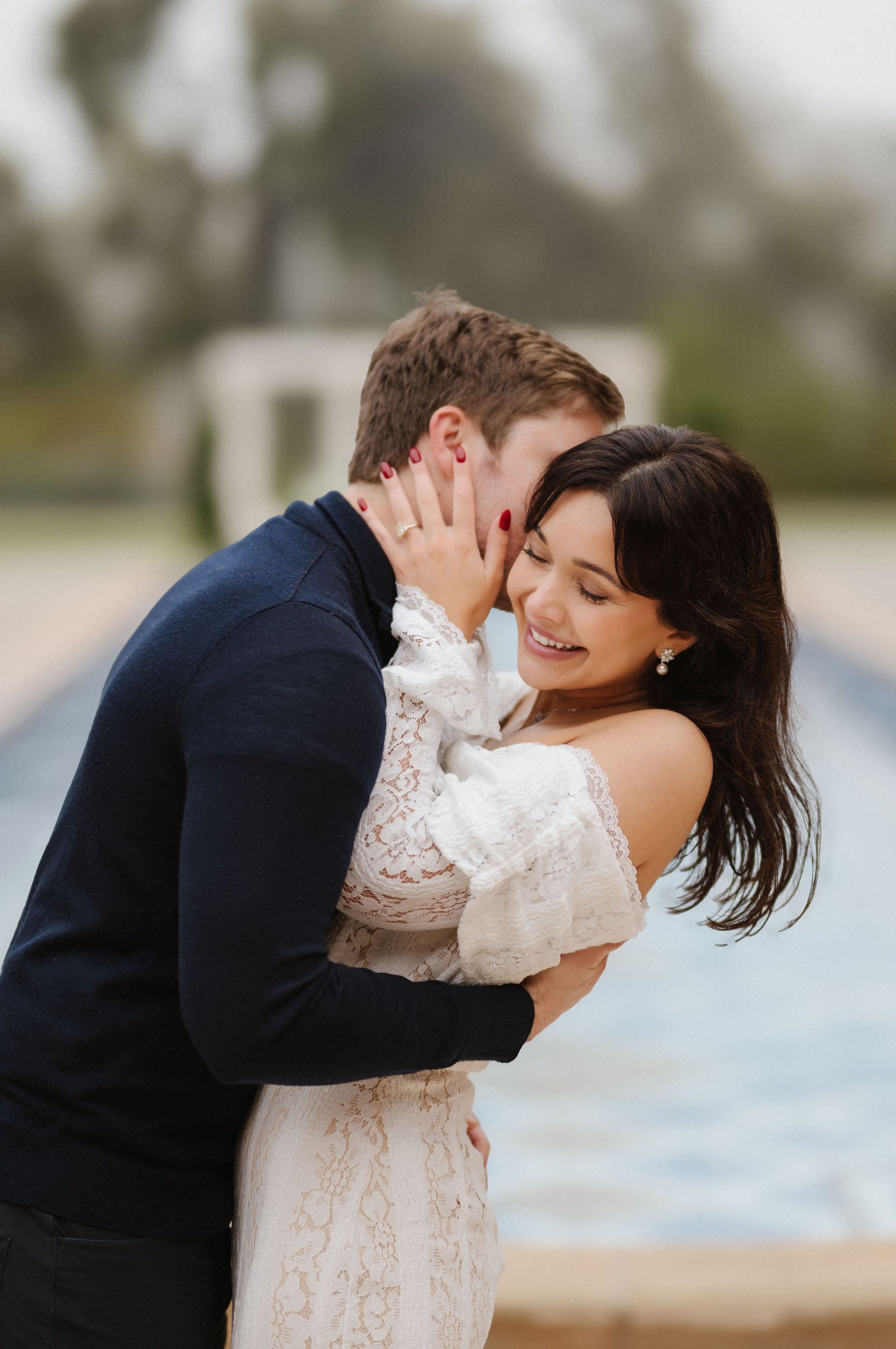 Couple embracing by a fountain for a proposal with The Yes Girls, smiling with blurred trees in the background 