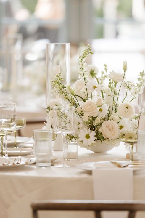 A photo with timeless editing of white floral centerpiece on a set dining table  in a soft, bright room
