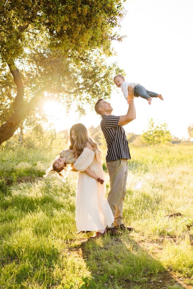 Family in a sunlit meadow, with one adult lifting a child and another holding a child near a tree.
