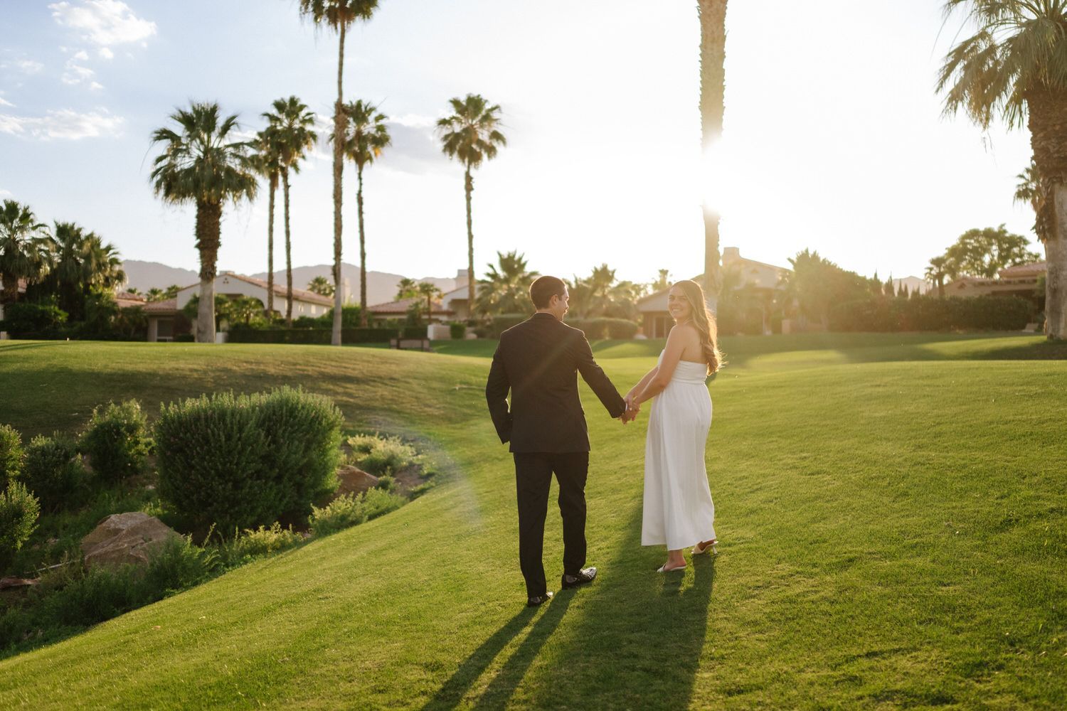 Couple holding hands while walking on a sunlit lawn with palm trees at sunset in Palm Desert, CA