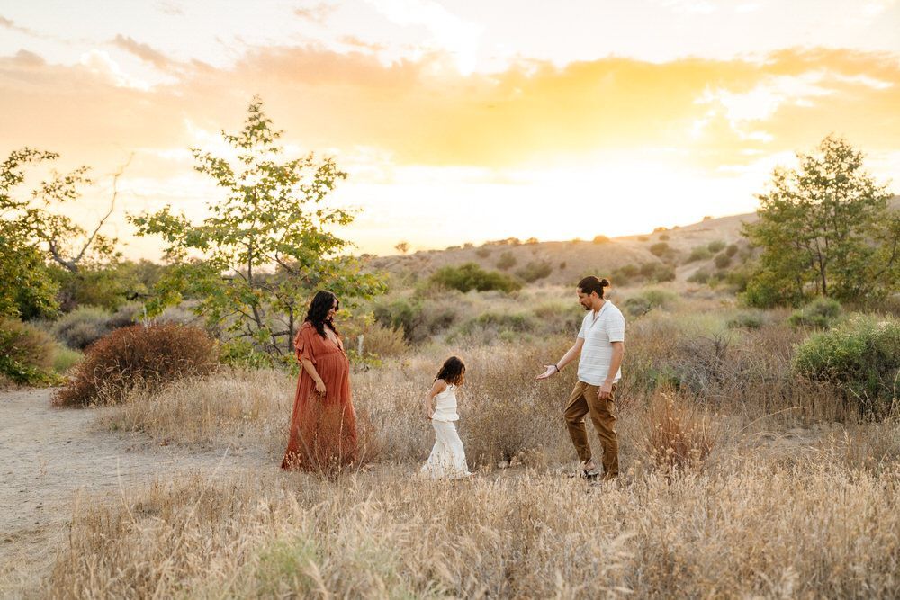 Three people walking through a dry field at sunset, with trees and golden sky in the background.